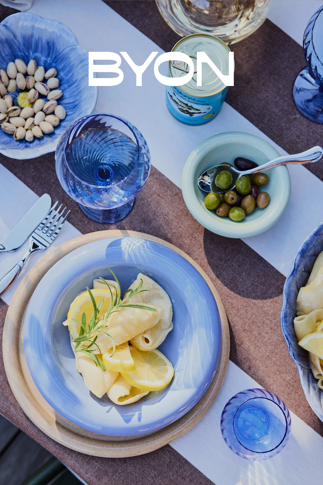 Vista aérea de una mesa de comedor con vajilla azul, incluyendo un plato de pasta con limón y romero, cuencos de pistachos y aceitunas, y vasos azules. El logotipo de BYON está superpuesto.