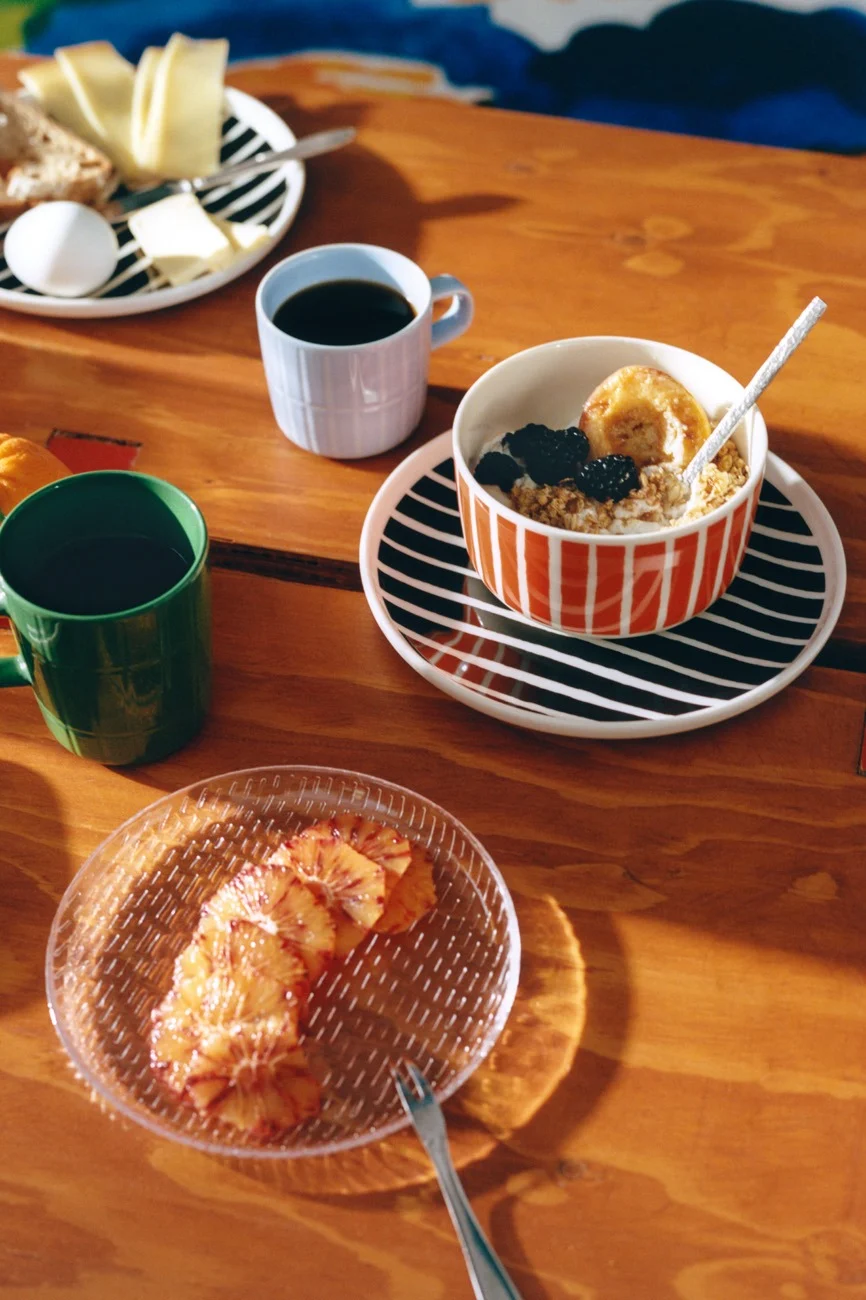 Desayuno en mesa de madera con vajilla a rayas, café, avena, queso, pan, huevo y rodajas de naranja.