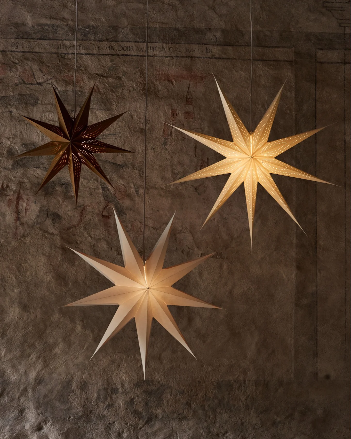 Tres farolillos de papel en forma de estrella cuelgan de una pared texturizada. Uno oscuro tiene pequeñas perforaciones, dos más brillantes iluminan la escena.