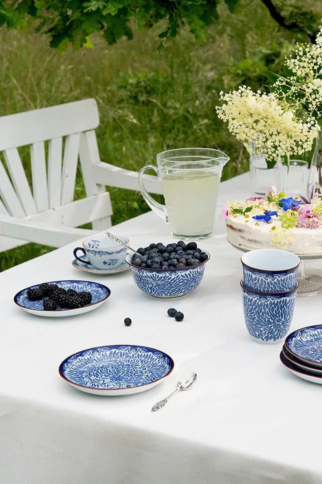 Mesa al aire libre con vajilla azul y blanca, arándanos y moras frescas, jarra de limonada y pastel con flores.