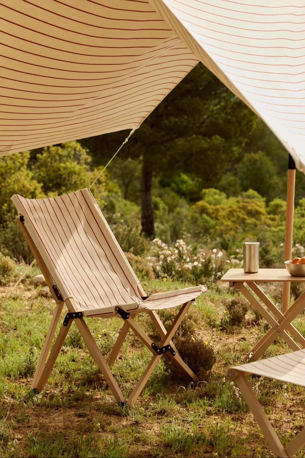 Un toldo a rayas dando sombra a una silla plegable de madera a rayas y a una mesa con un termo y unas naranjas al aire libre.