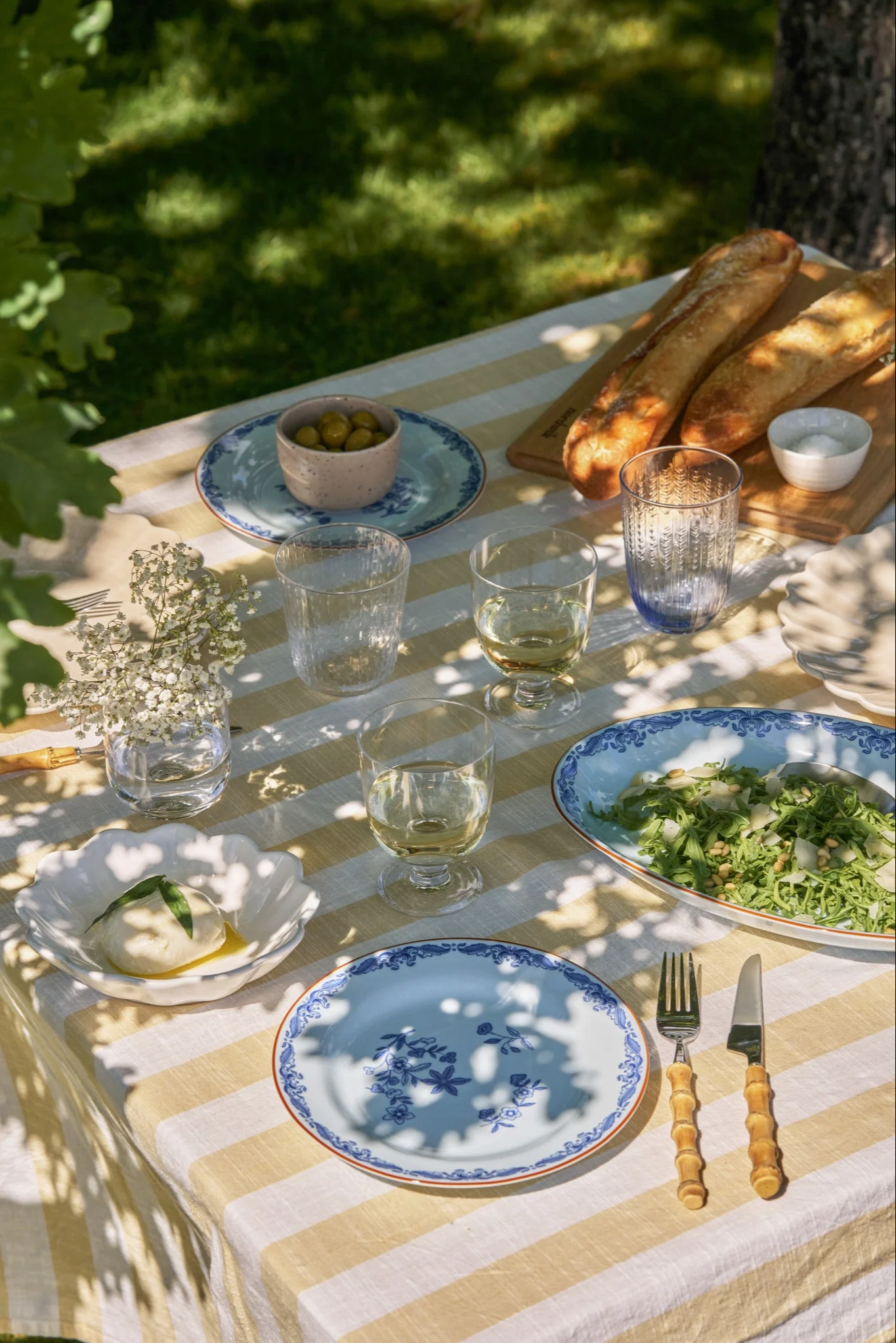 Una mesa de picnic al aire libre con mantel de rayas amarillas y blancas, burrata, ensalada, baguettes, aceitunas y platos azules.