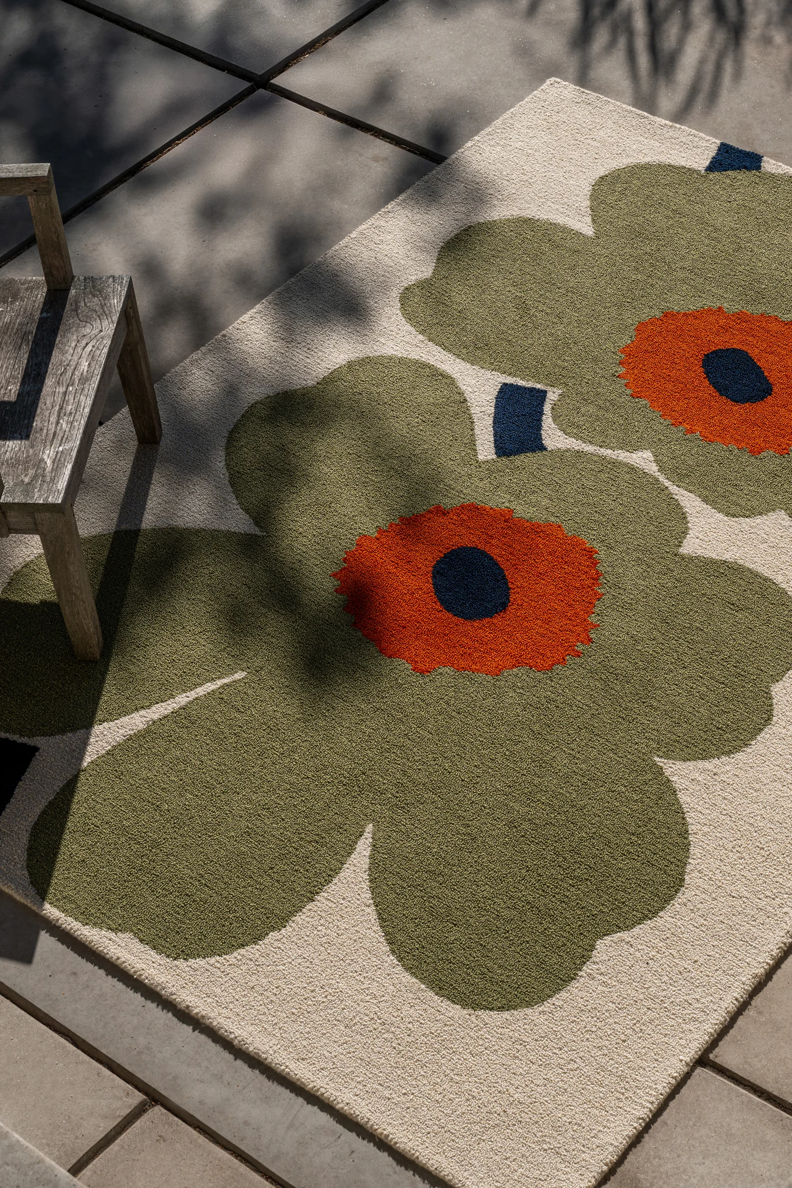 Alfombra con grandes motivos florales verdes y naranjas sobre fondo claro, bajo una silla de madera en patio, con sombras de hojas.