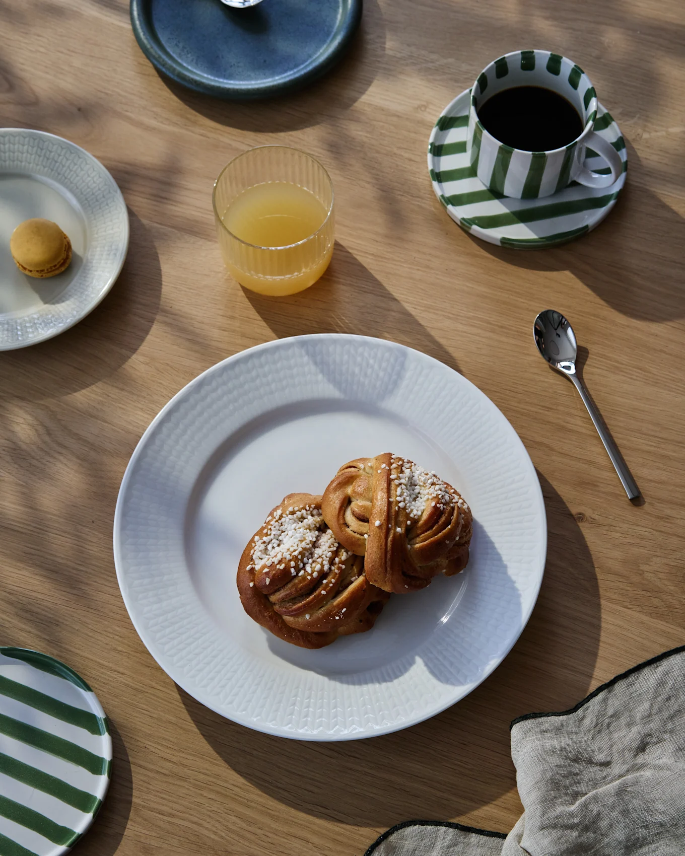 Un desayuno en una mesa de madera con dos rollos de canela, café en una taza a rayas, zumo de naranja y un macaron.