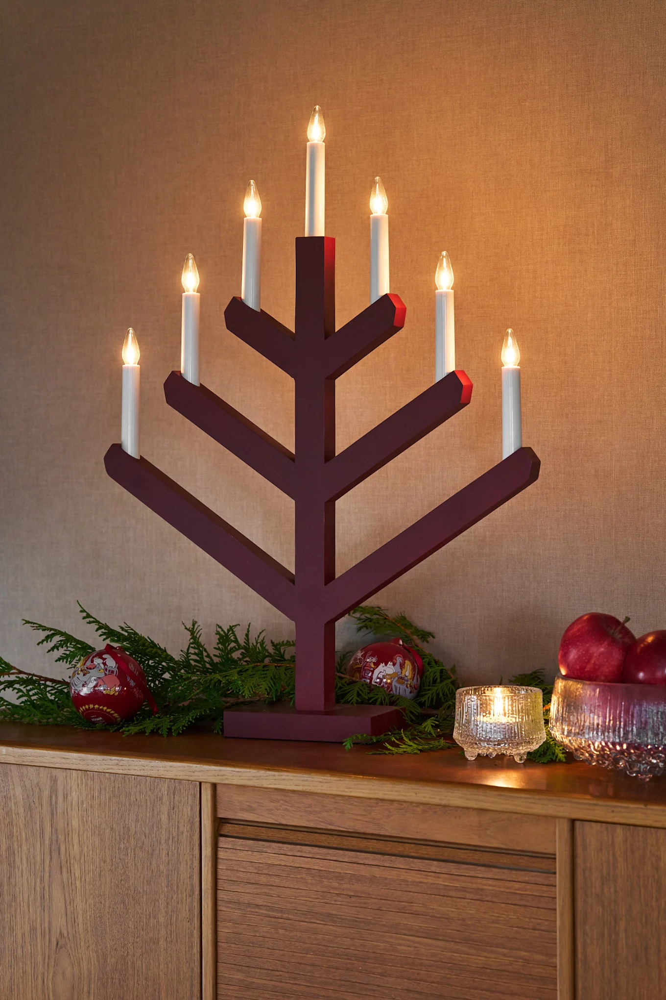 El candelabro Pine de Scandi Living está sobre una cómoda junto con una linterna y un cuenco Ultima Thule de Iittala lleno de manzanas. Bolas de Navidad y ramitas de abeto completan la decoración festiva.