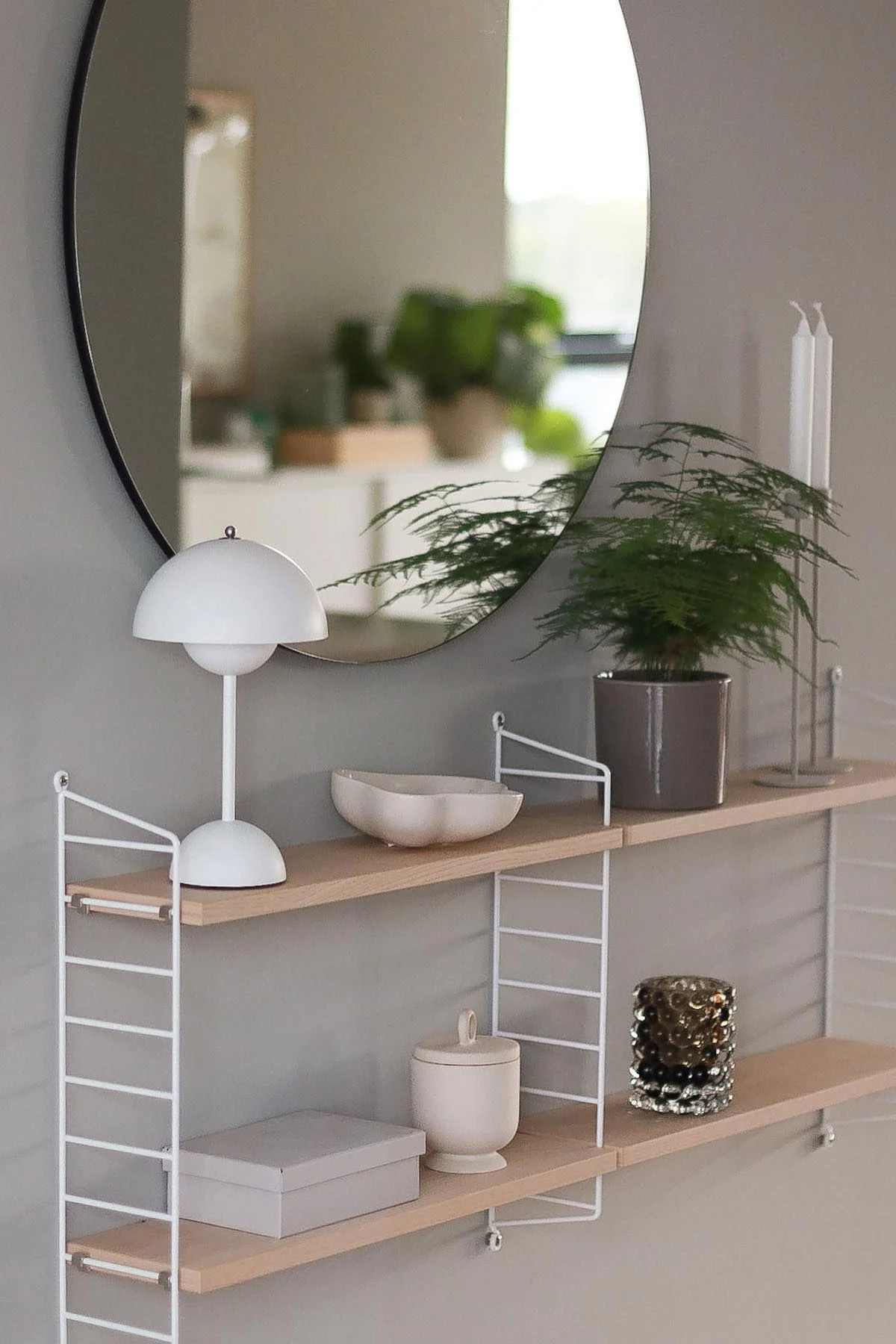 Un espejo redondo cuelga sobre estantes de pared blancos con una lámpara blanca de seta, una planta de helecho, objetos y velas.