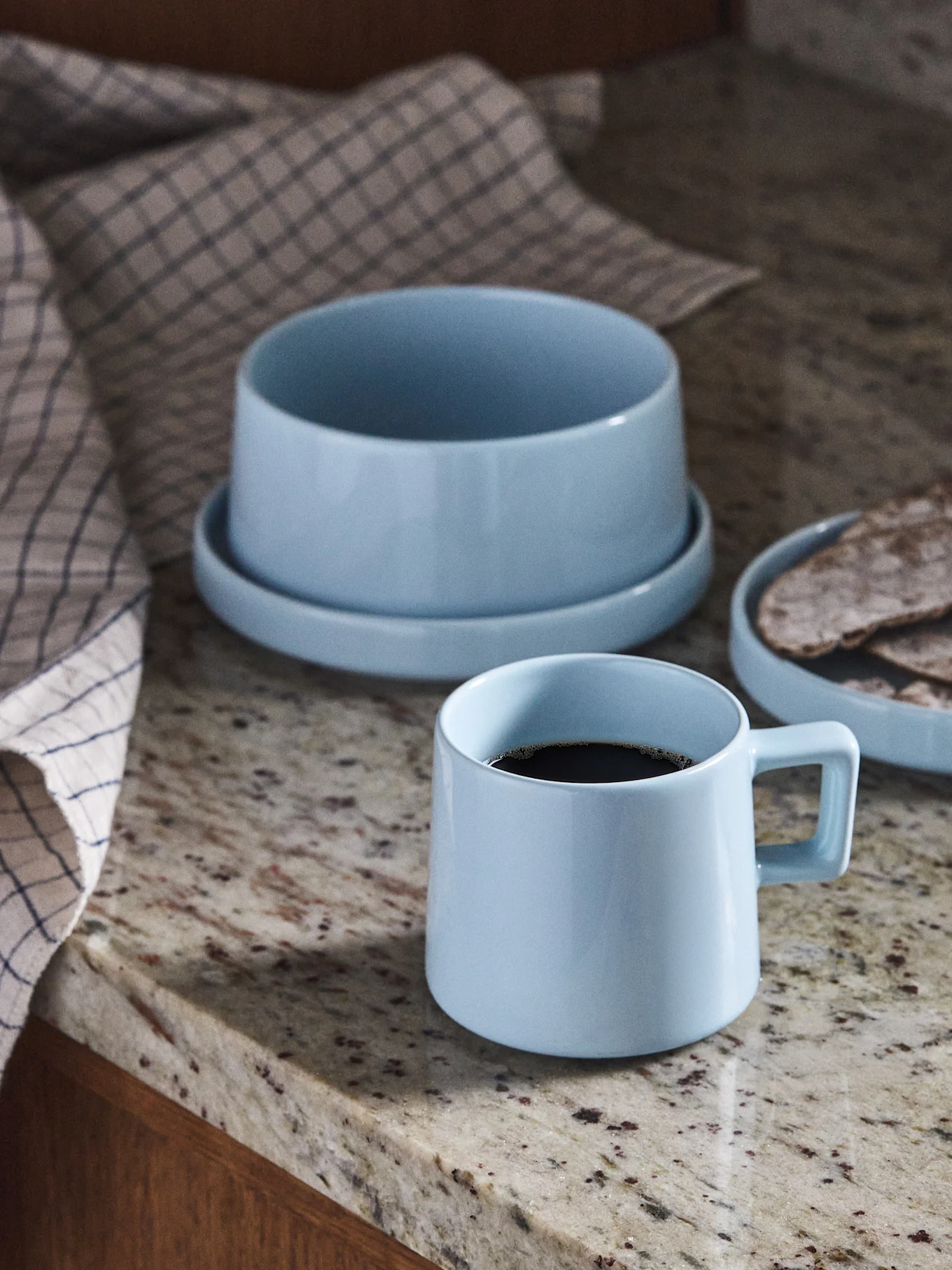 Taza de café de cerámica azul claro, cuenco y plato con pan plano sobre encimera de granito.