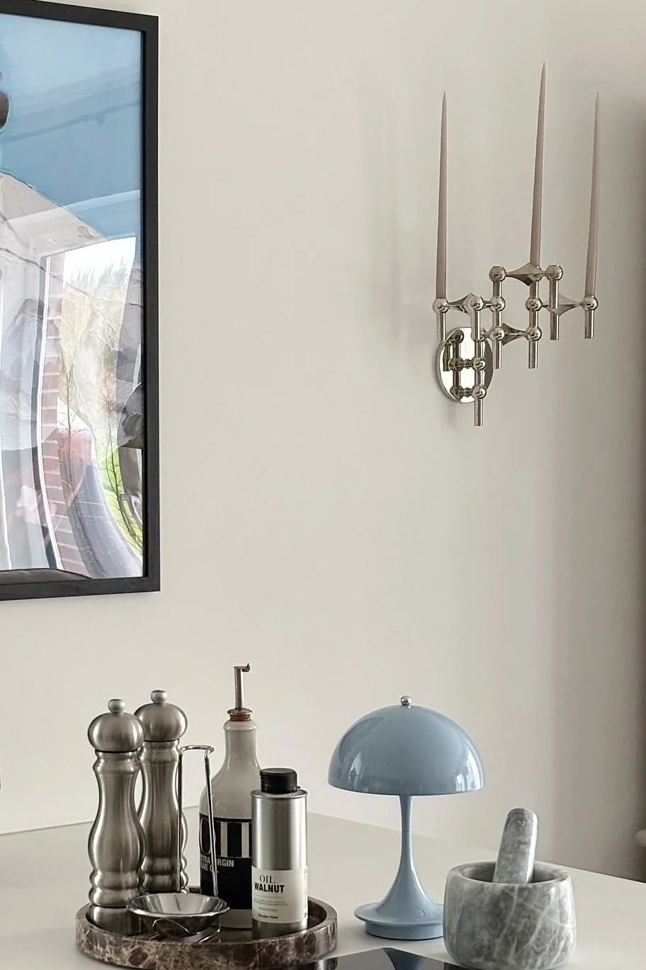 Un aplique de pared plateado con tres velas cuelga sobre una mesa blanca con artículos de cocina: molinillos, botellas de aceite, una lámpara azul de seta y un mortero de mármol.