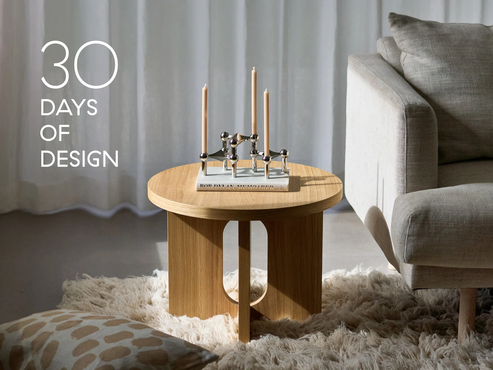Sala de estar moderna con mesa auxiliar redonda de madera, candelabro plateado con tres velas, un libro y un sofá beige. Texto: '30 DÍAS DE DISEÑO'.