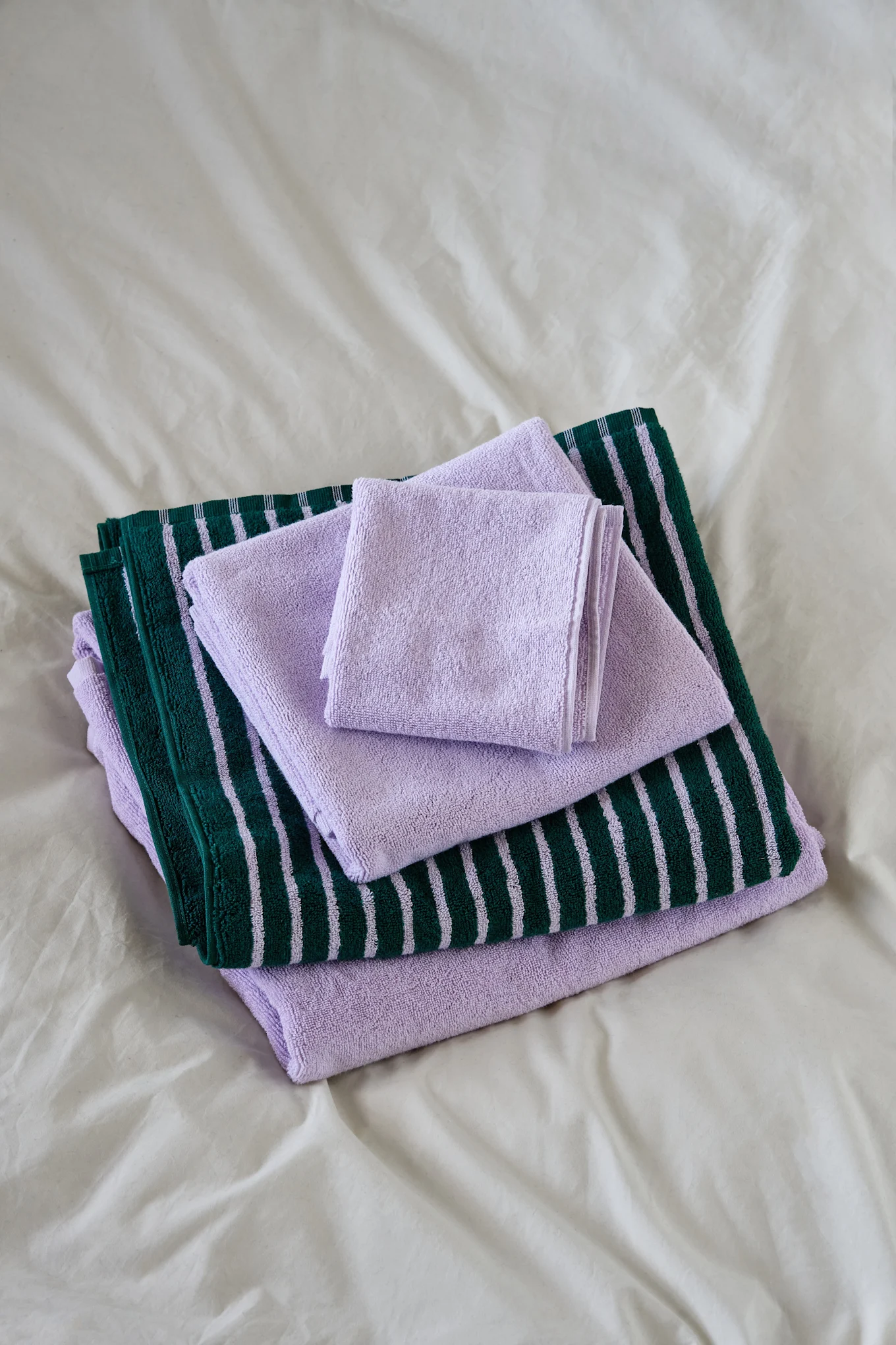 Toallas plegadas apiladas: morado claro y verde oscuro con rayas morado claro, sobre una sábana blanca.