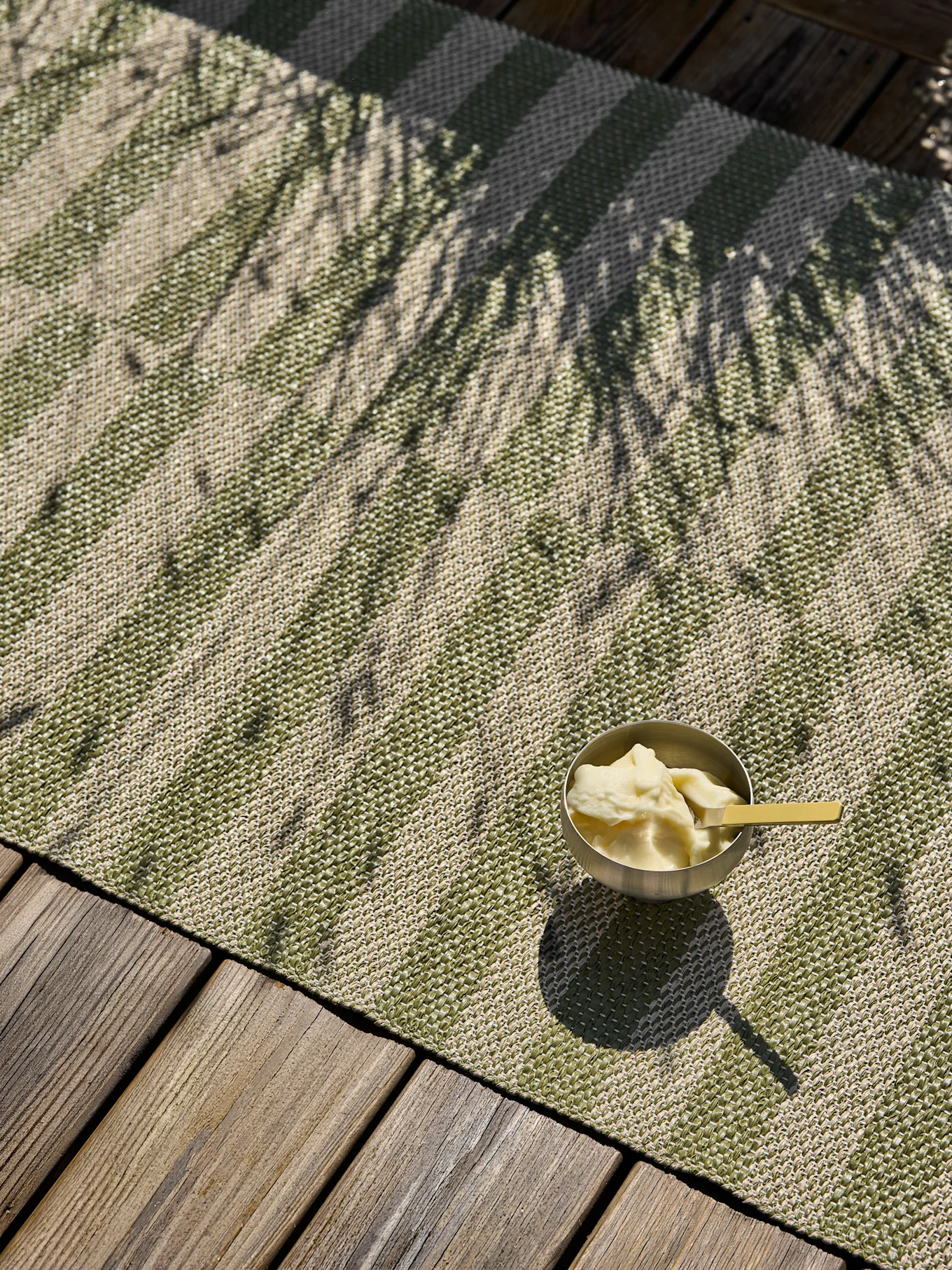 Un cuenco plateado con postre y una cuchara amarilla reposa sobre una alfombra exterior a rayas verdes y beige en una cubierta de madera, salpicada por sombras de palmeras.