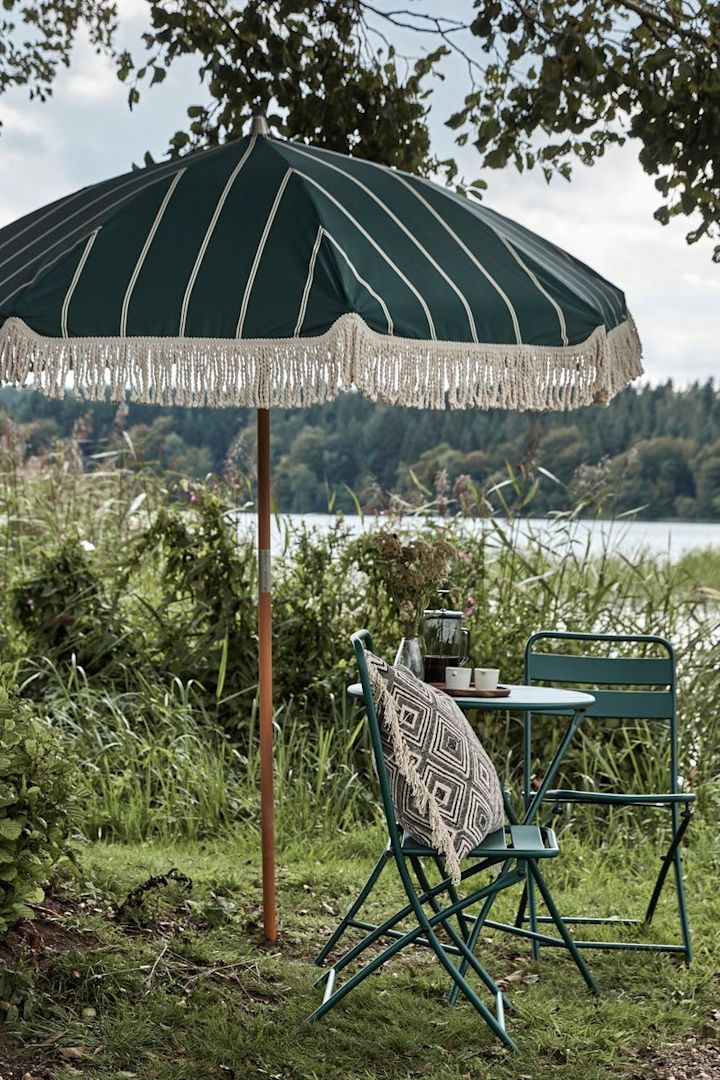 Decoración boho para el jardín: la sombrilla de bloque verde con flecos de House Doctor en un jardín, con una pequeña zona de asientos al lado.