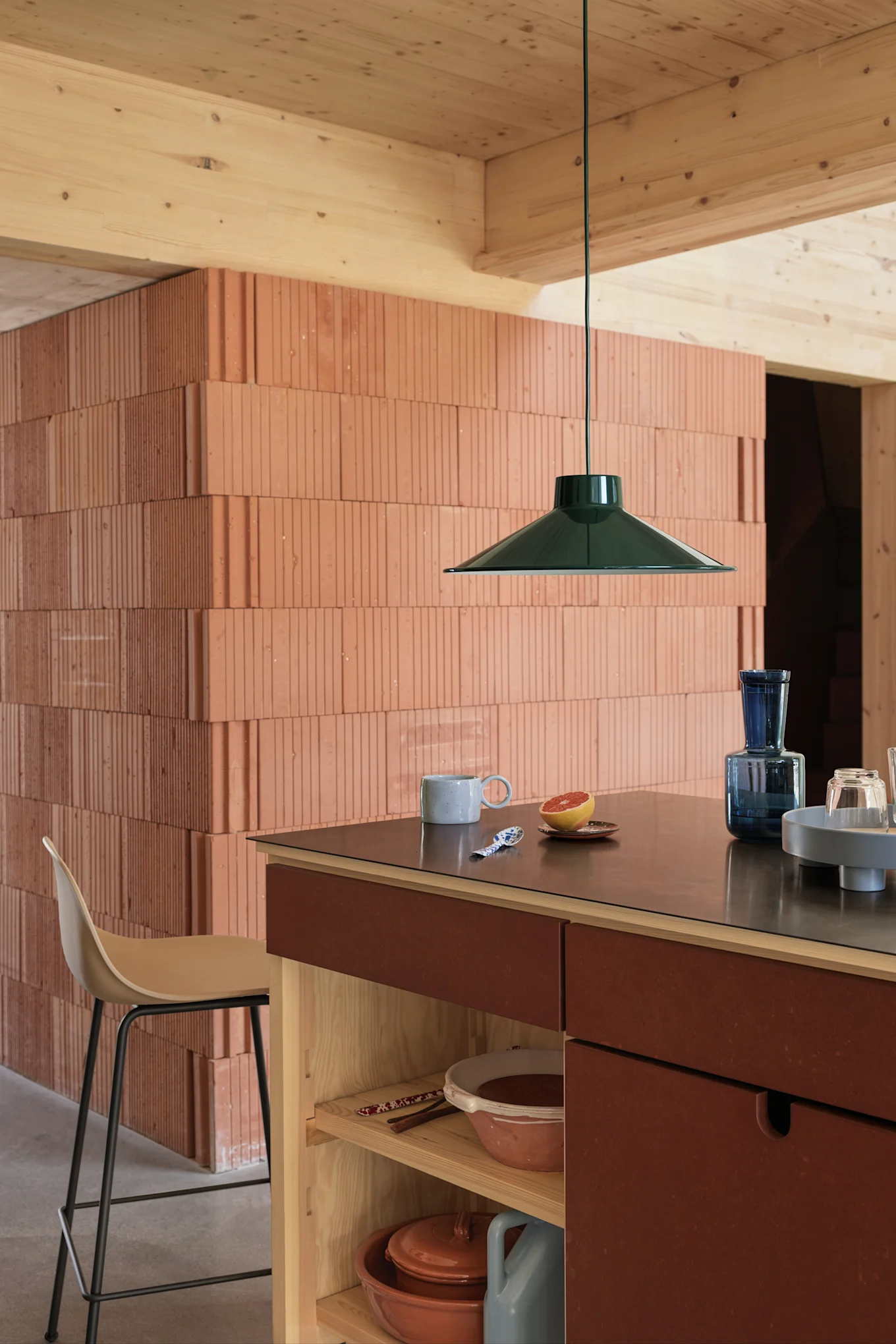 Lámpara colgante verde oscuro sobre una isla de cocina con encimera oscura, taza, pomelo y cristalería azul, pared de ladrillo texturizado.