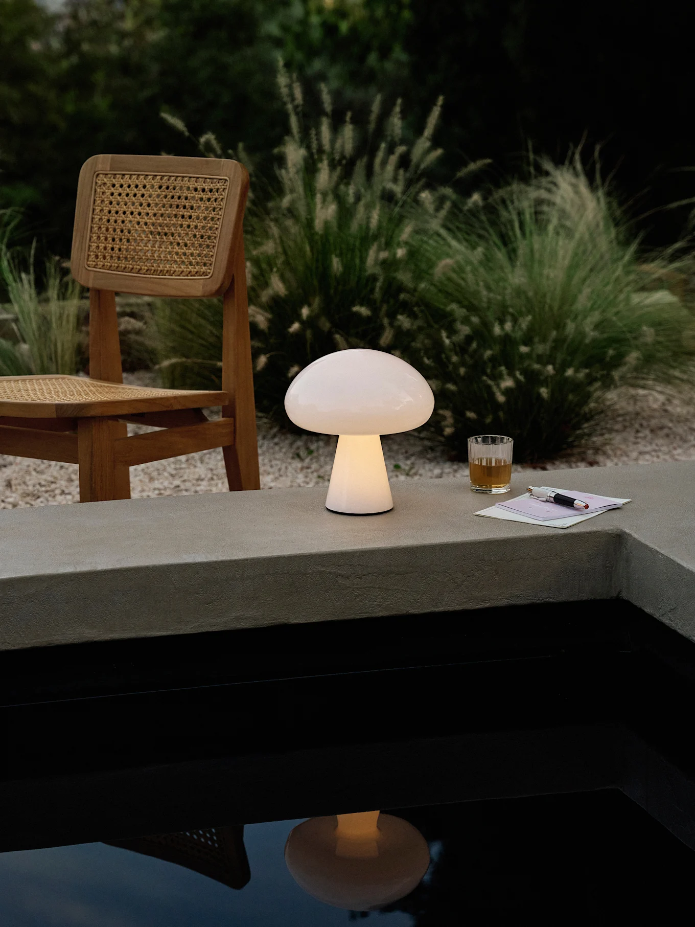 Una lámpara brillante con forma de seta sobre un saliente de hormigón junto a una piscina, con una silla de madera, bebida y bolígrafo.