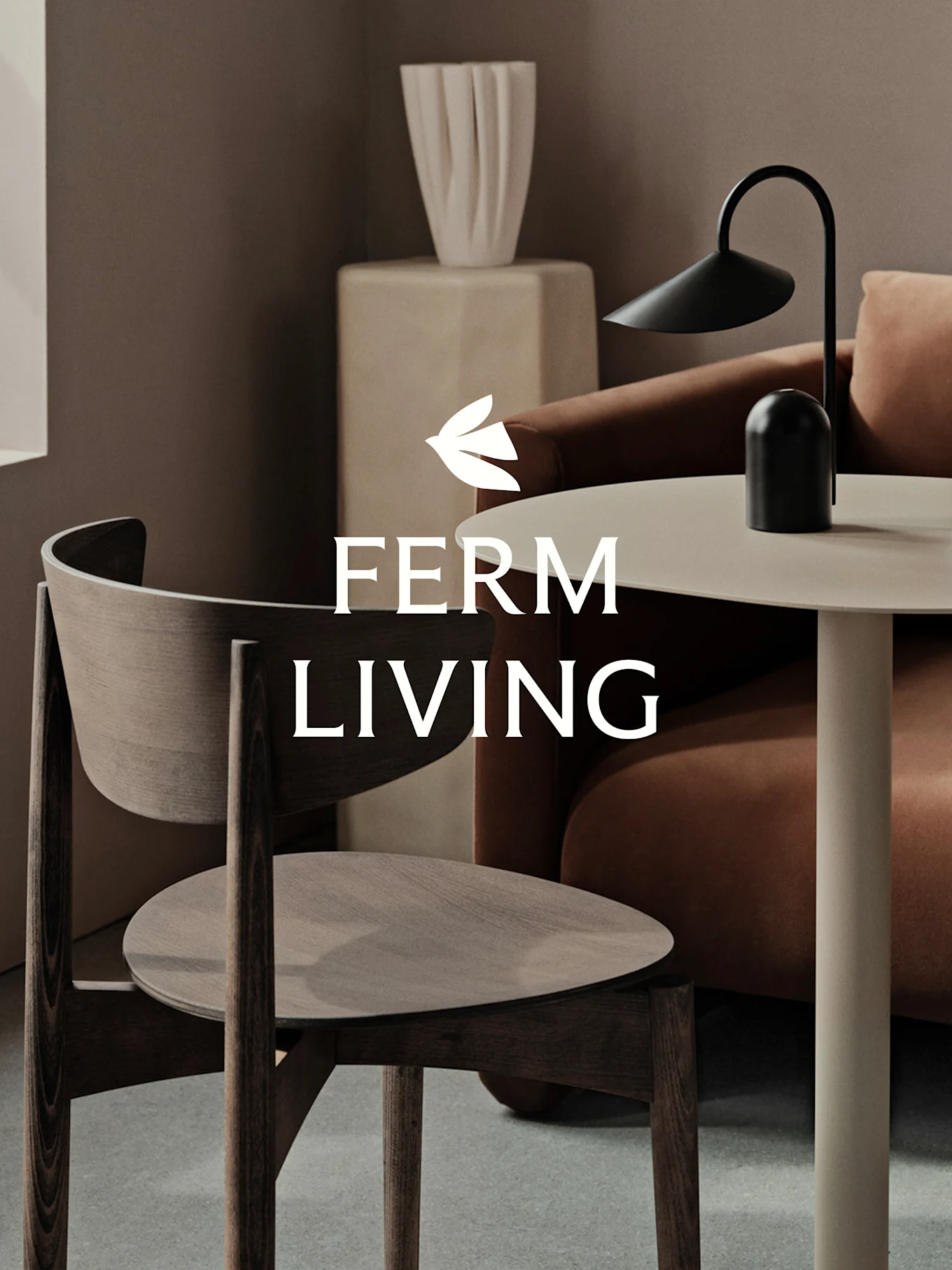 Un espacio moderno con una silla de madera oscura, una mesa auxiliar clara con una lámpara negra, un sofá terracota y un jarrón escultural. Logo 'FERM LIVING'.