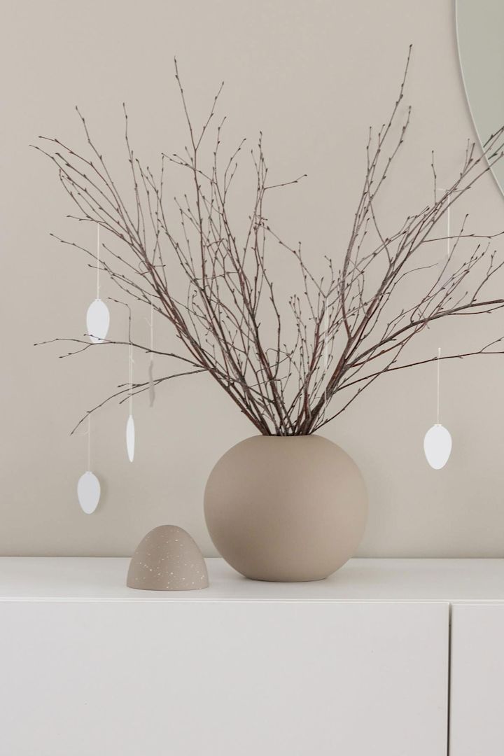 Un arbusto de Pascua también se encuentra sobre una cómoda blanca en un jarrón esférico de color arena con colgantes de huevo de Pascua, ambos de Cooee Design, en el hogar de @tunadalen.