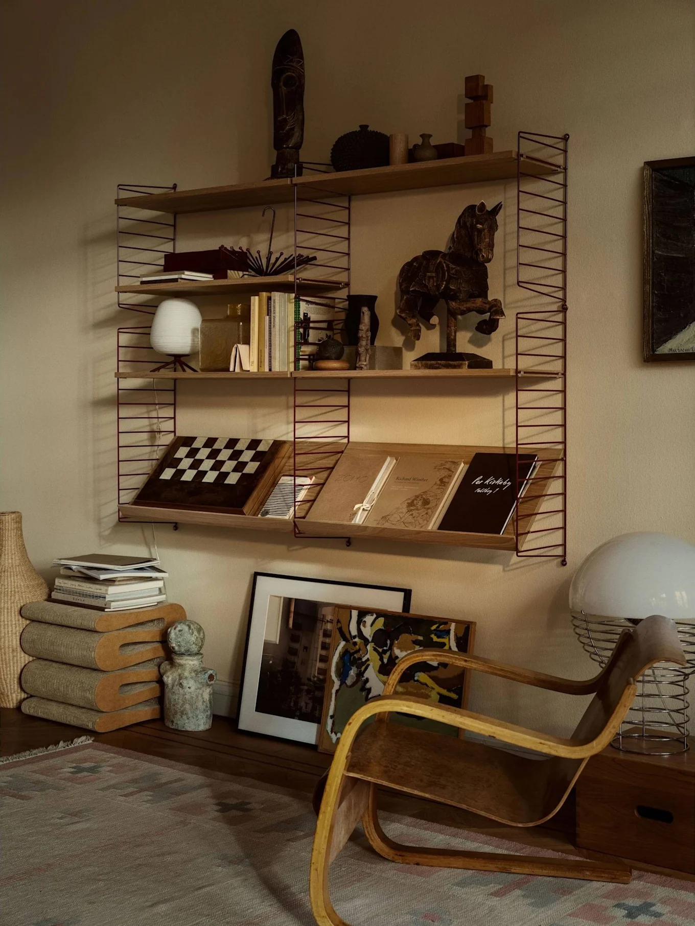 Una acogedora habitación con una estantería llena de libros, arte y decoración, junto a un sillón de madera curvada y una lámpara blanca grande.