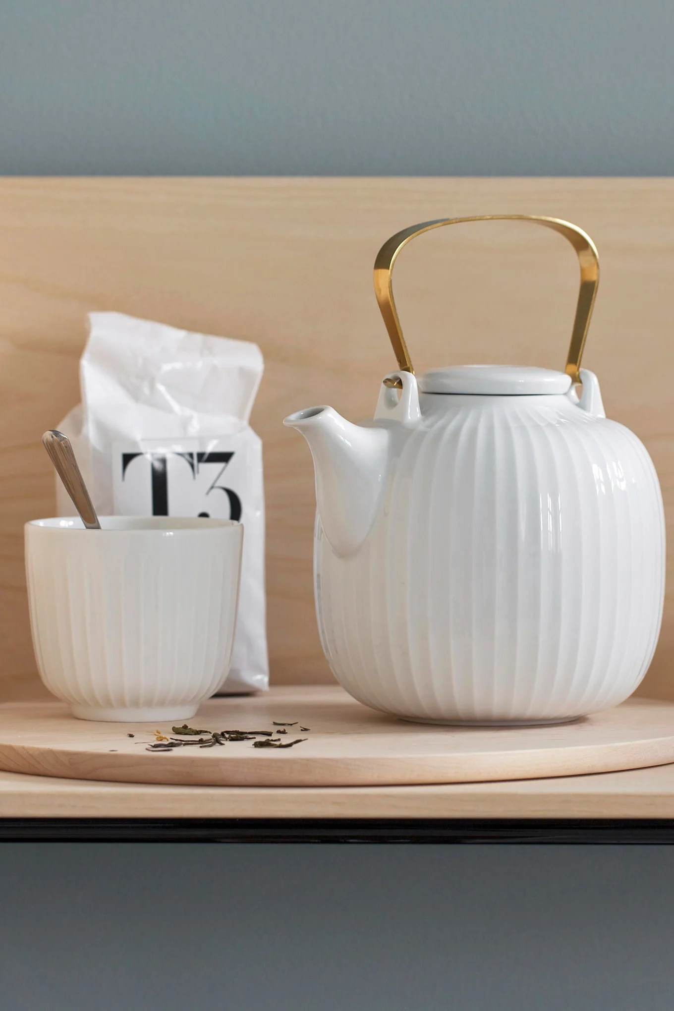 Tetera blanca acanalada con asa dorada, taza a juego con cuchara y una bolsa de té en un estante de madera.