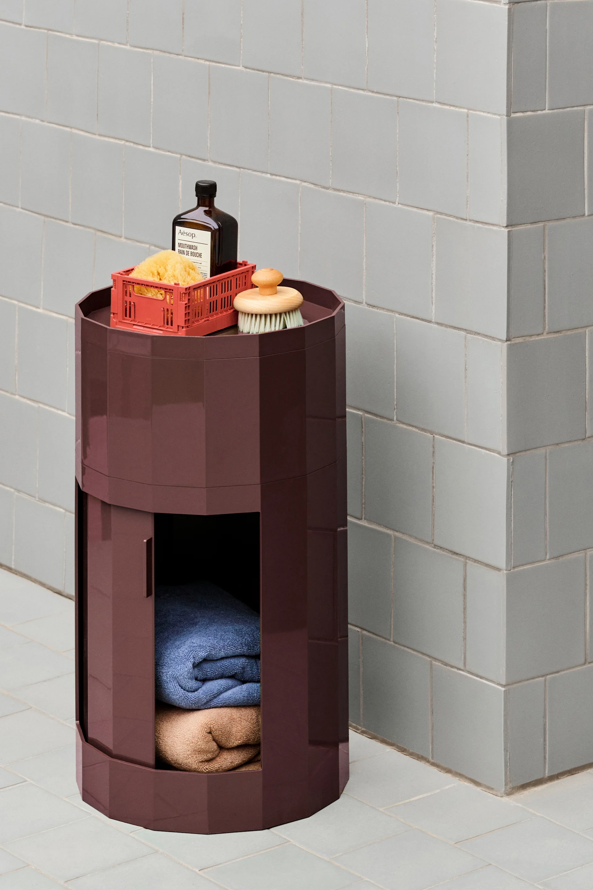 Un mueble de almacenamiento hexagonal rojo oscuro con productos de baño encima y toallas dobladas dentro, sobre una pared de azulejos gris claro.