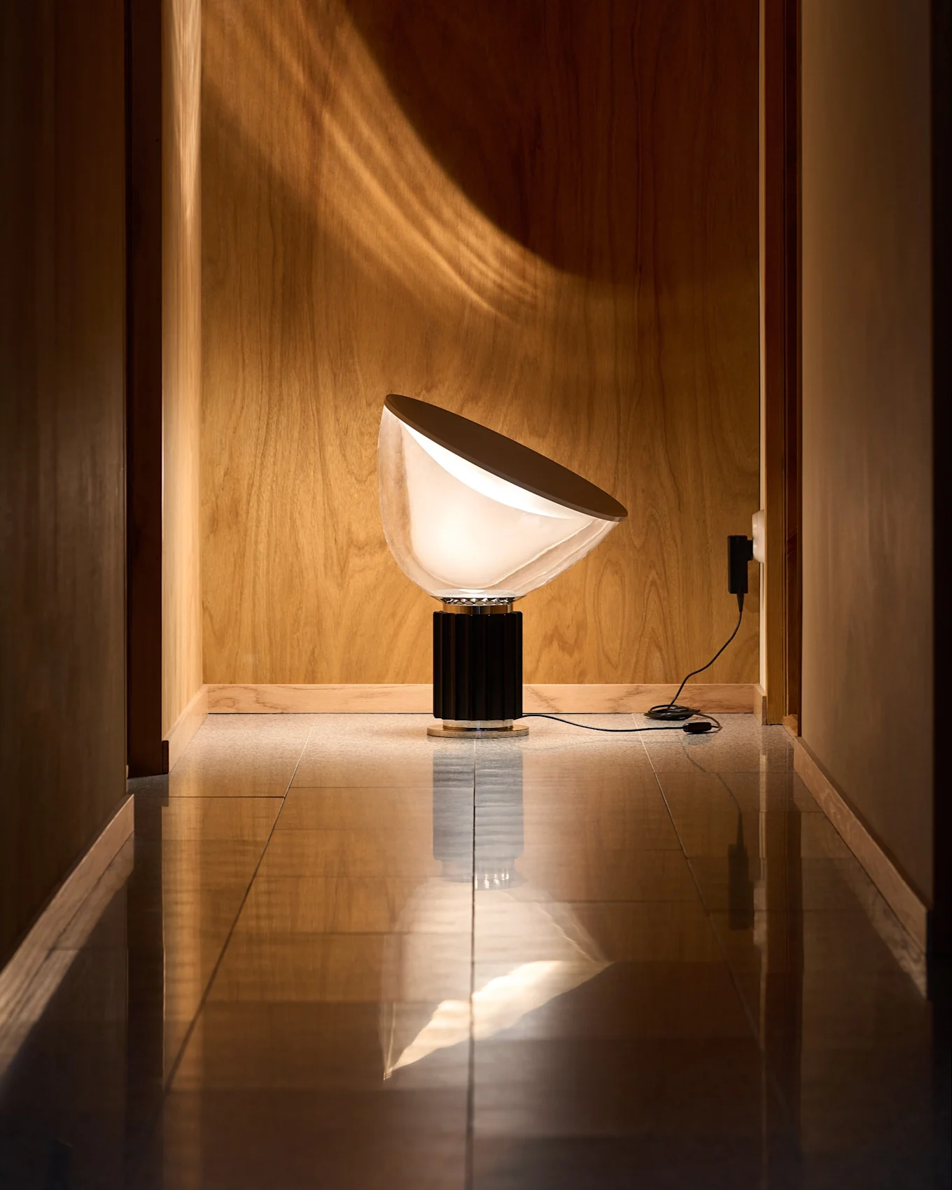 Lámpara moderna con pantalla semicircular blanca opaca y base negra estriada en pasillo de madera con suelo reflectante. Luz proyecta patrón curvo.