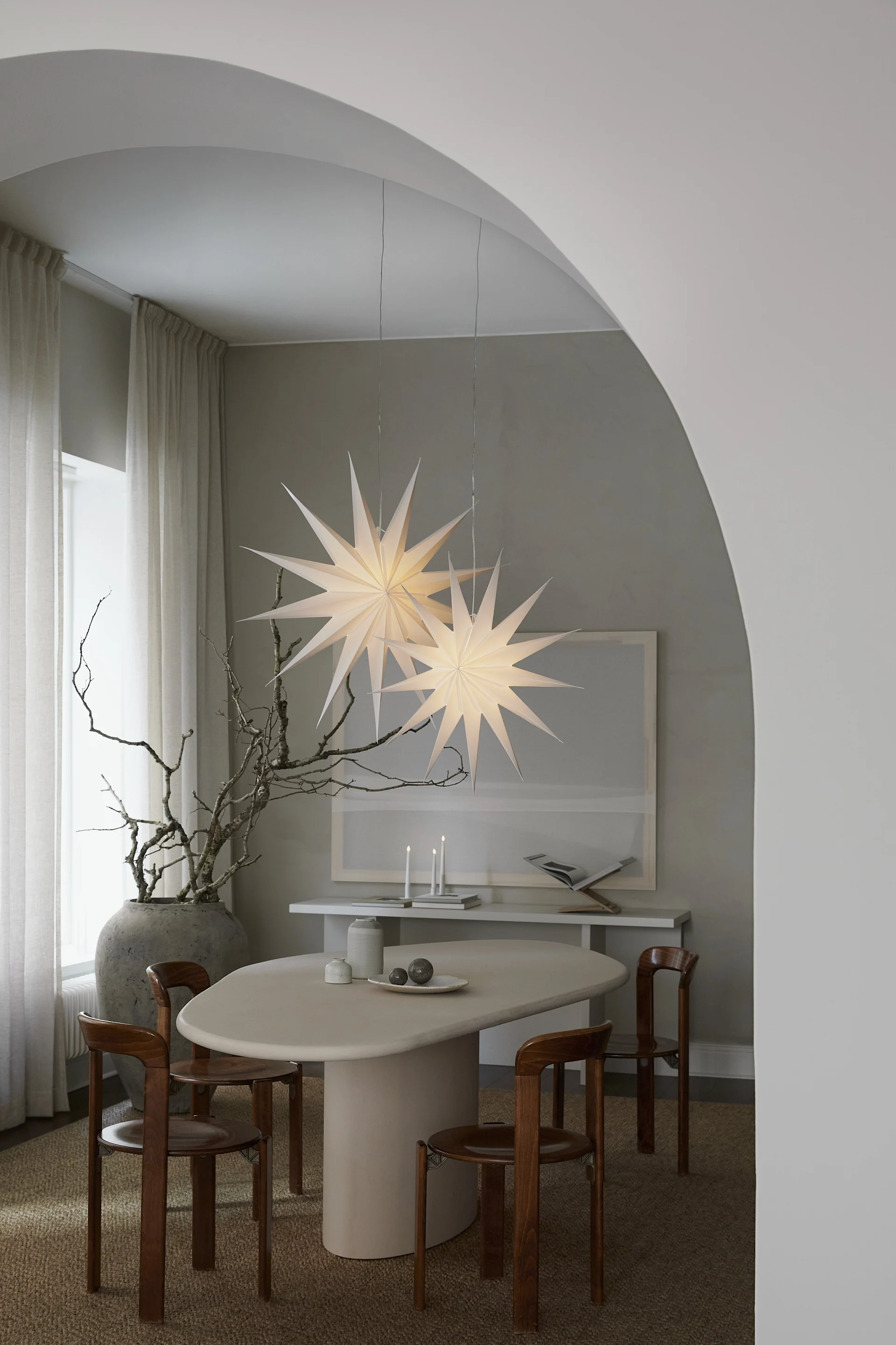 Comedor moderno con mesa ovalada, sillas de madera, dos lámparas colgantes grandes en forma de estrella y ramas secas en un jarrón.
