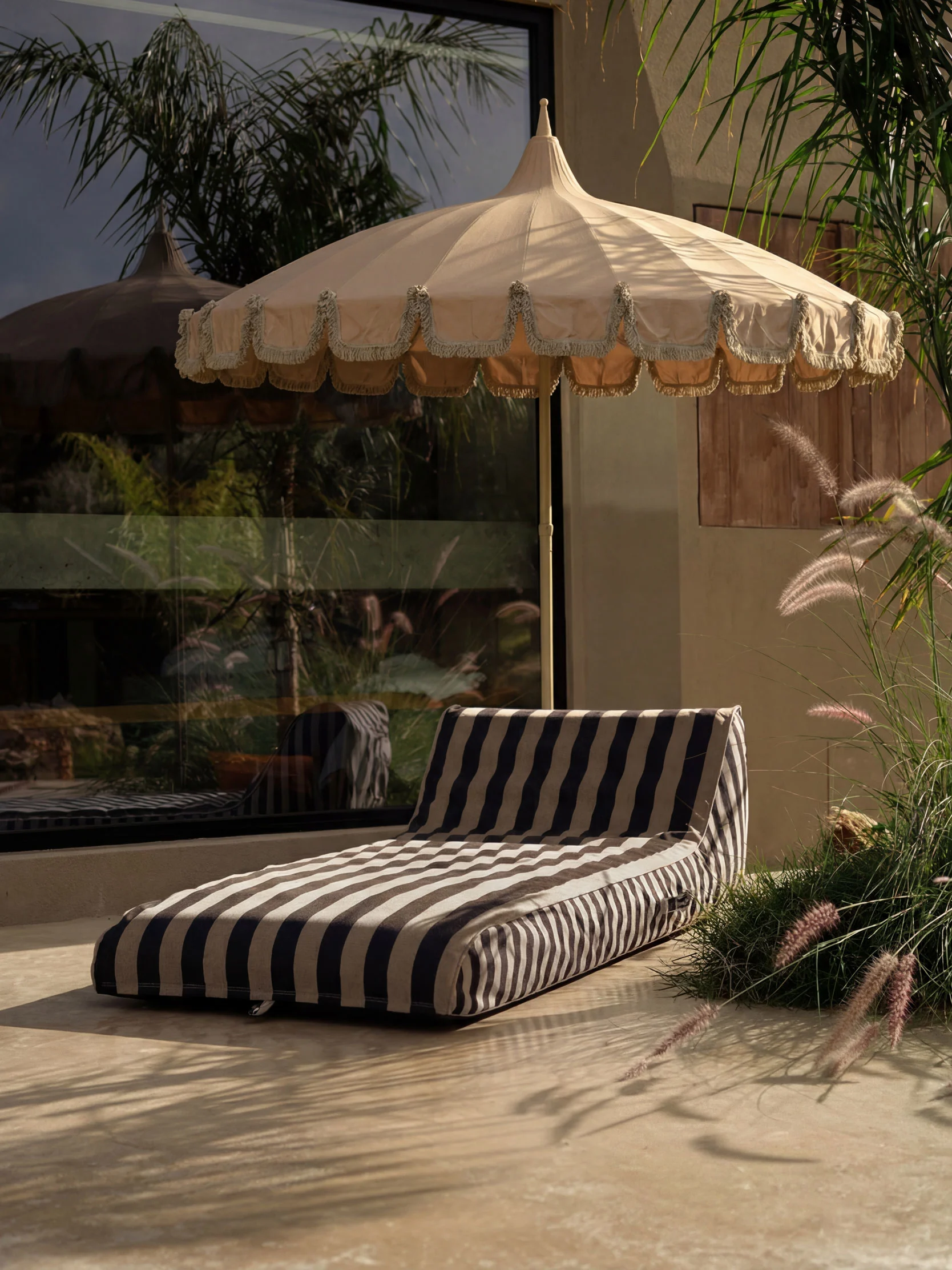 Patio exterior con sombrilla beige con flecos y cojín de rayas en el suelo claro, rodeado de plantas.