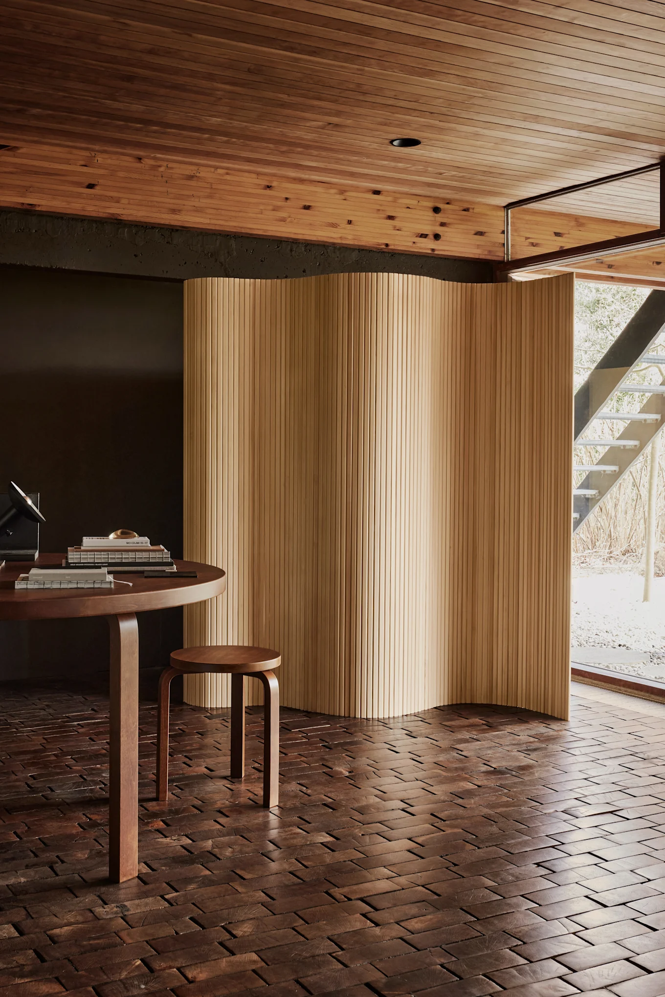 Habitación moderna con mesa redonda de madera, un taburete pequeño y una pantalla de madera ondulada. Suelo de bloques de madera oscura, techo de listones de madera clara y una ventana a una escalera exterior.
