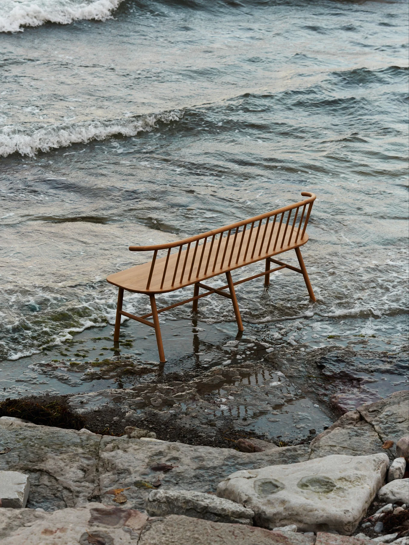 Un banco de madera clara con listones y respaldo curvo se encuentra en aguas poco profundas en una playa rocosa con olas suaves.