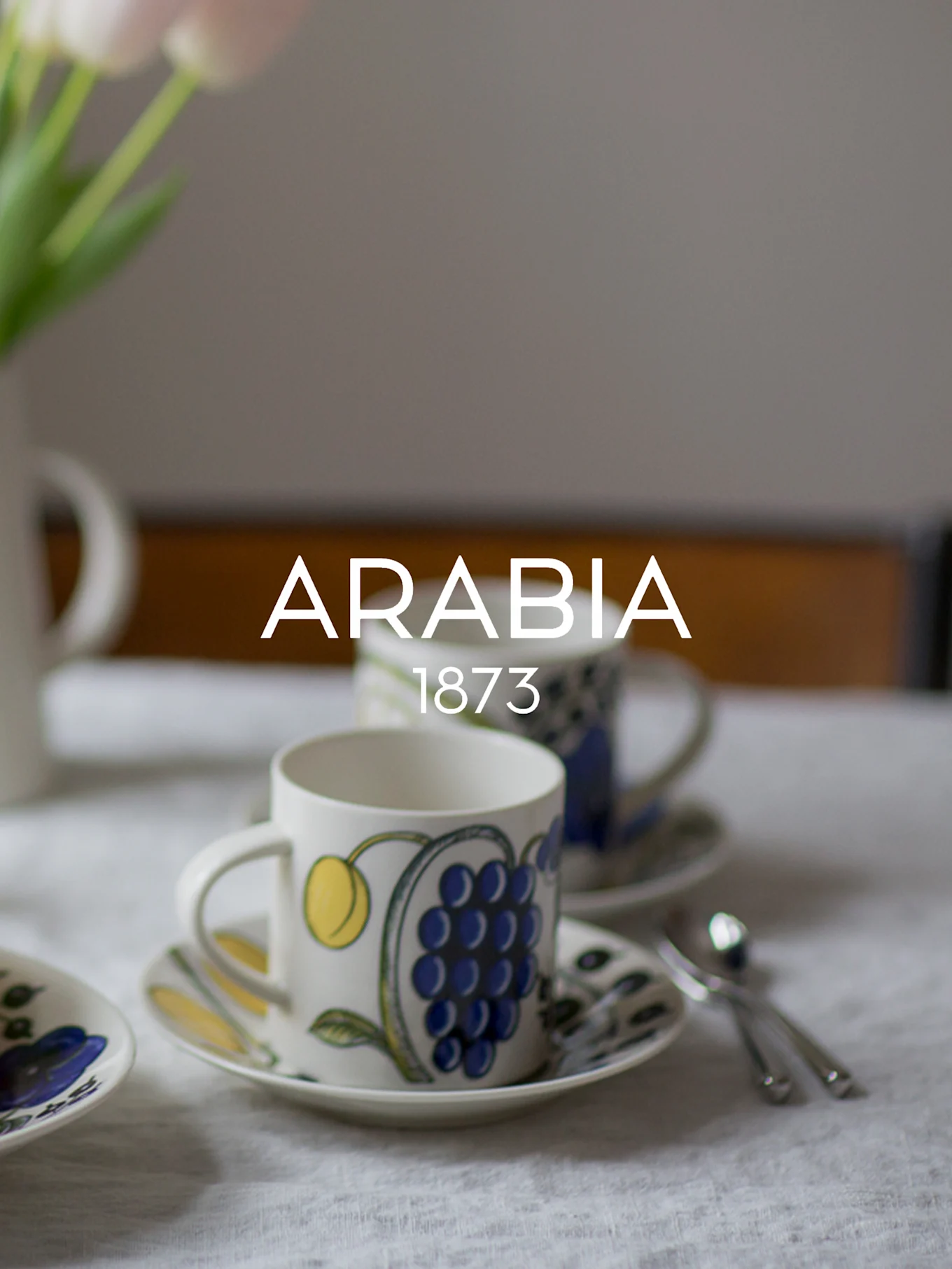 Dos tazas y platillos de cerámica estampados, uno con uvas azules, sobre un mantel blanco. El texto 'ARABIA 1873' está superpuesto.