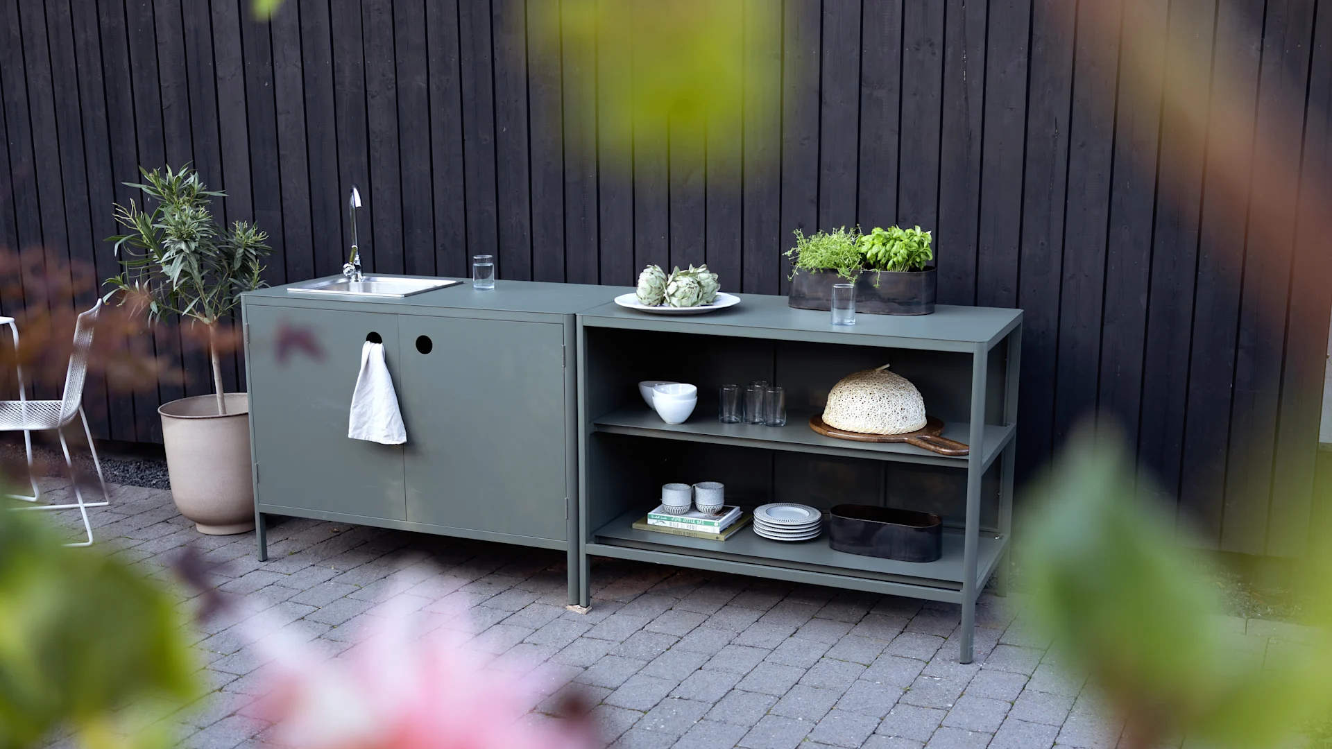 Mueble de cocina exterior verde oliva con fregadero, estantes abiertos, hierbas en macetas, vajilla y una campana de mimbre en un patio.