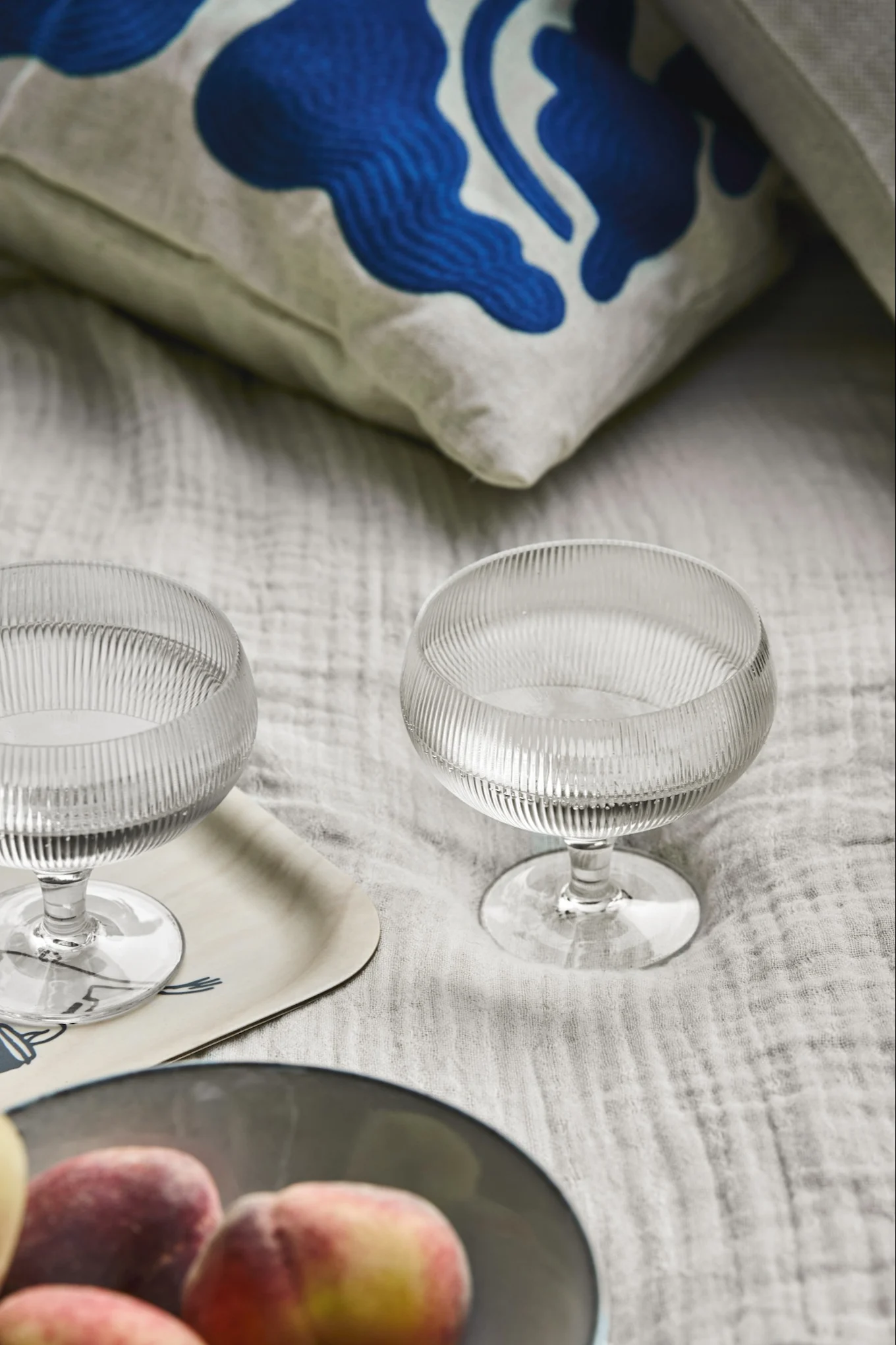 Dos copas estriadas de cristal, un cuenco de melocotones y un cojín estampado sobre textil claro.