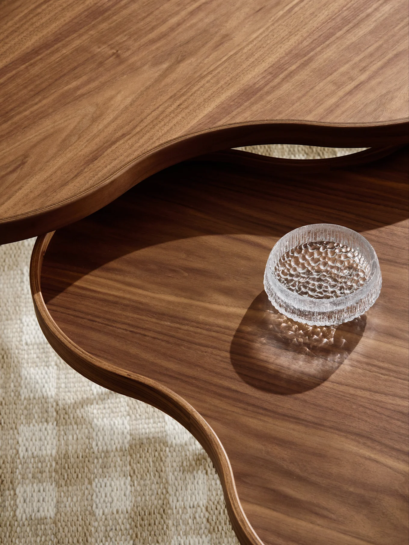 Primer plano de mesa de centro de madera de varios niveles con bordes ondulados y cuenco de cristal texturizado en nivel inferior, sobre alfombra.