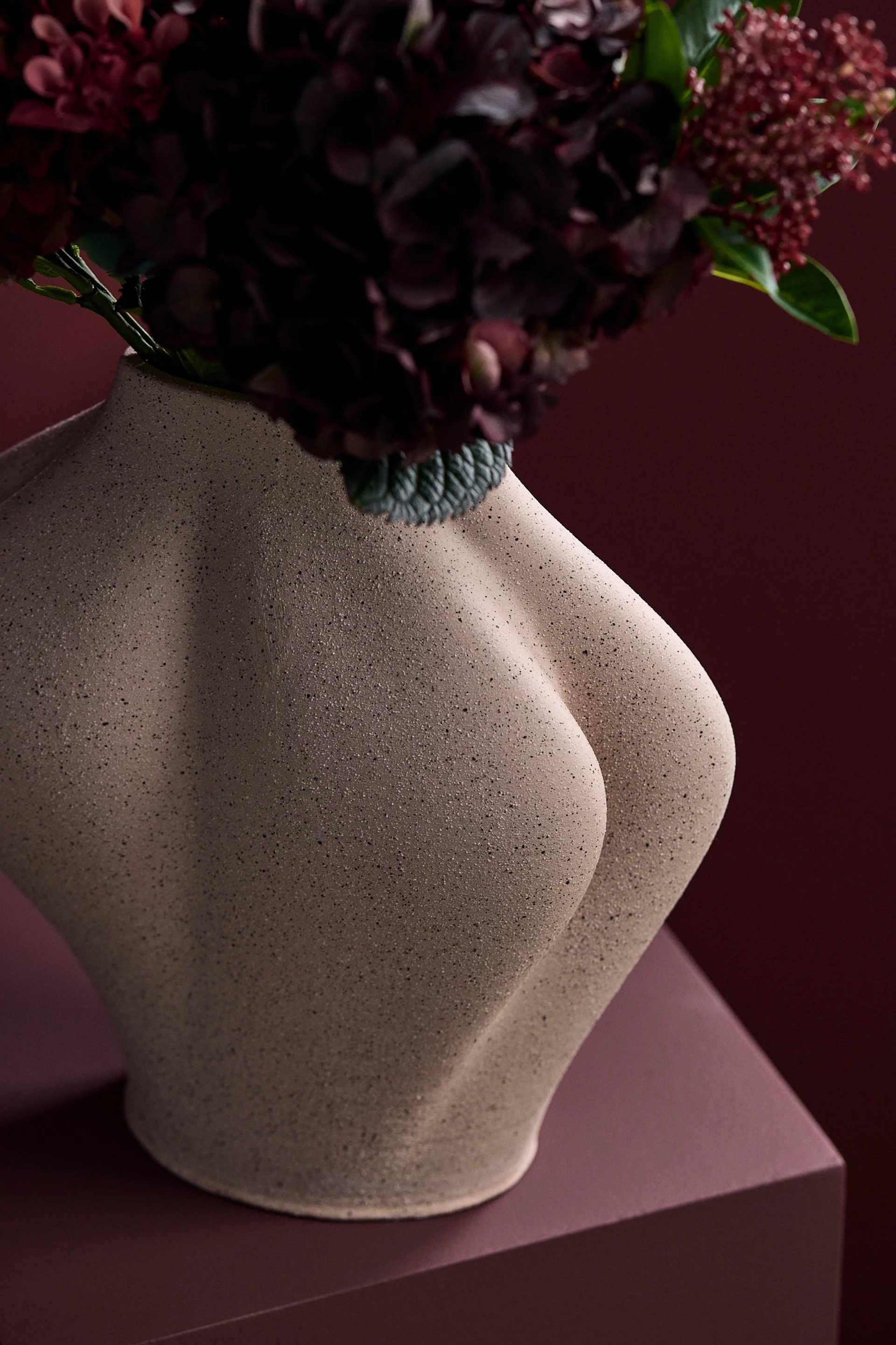 Jarrón escultural beige con forma de torso humano, con flores rojas oscuras sobre un fondo rojo a juego.