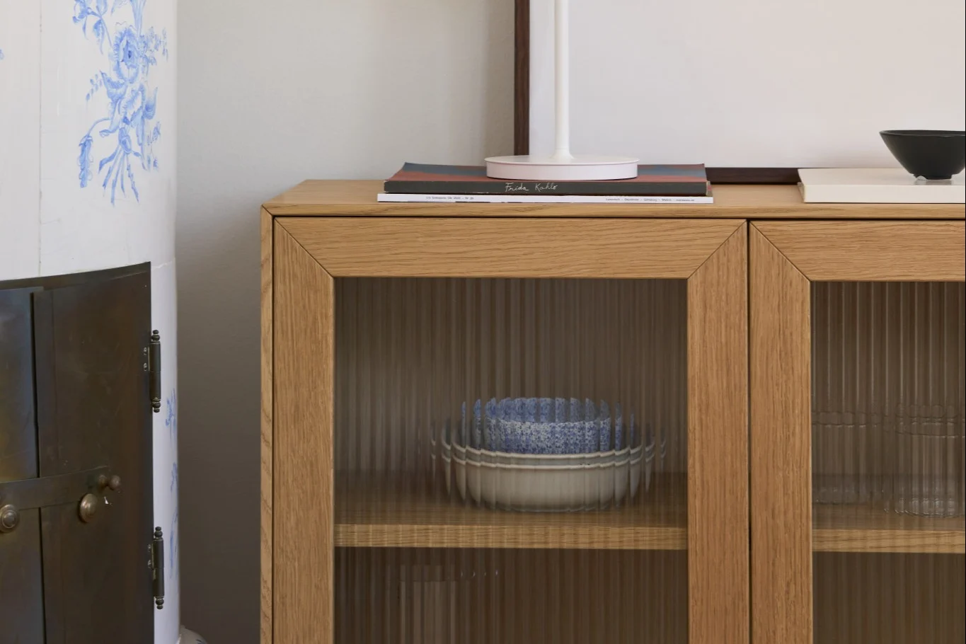 Gabinete de madera con puertas de vidrio estriado, con cuencos apilados azules y blancos, junto a una estufa de azulejos.