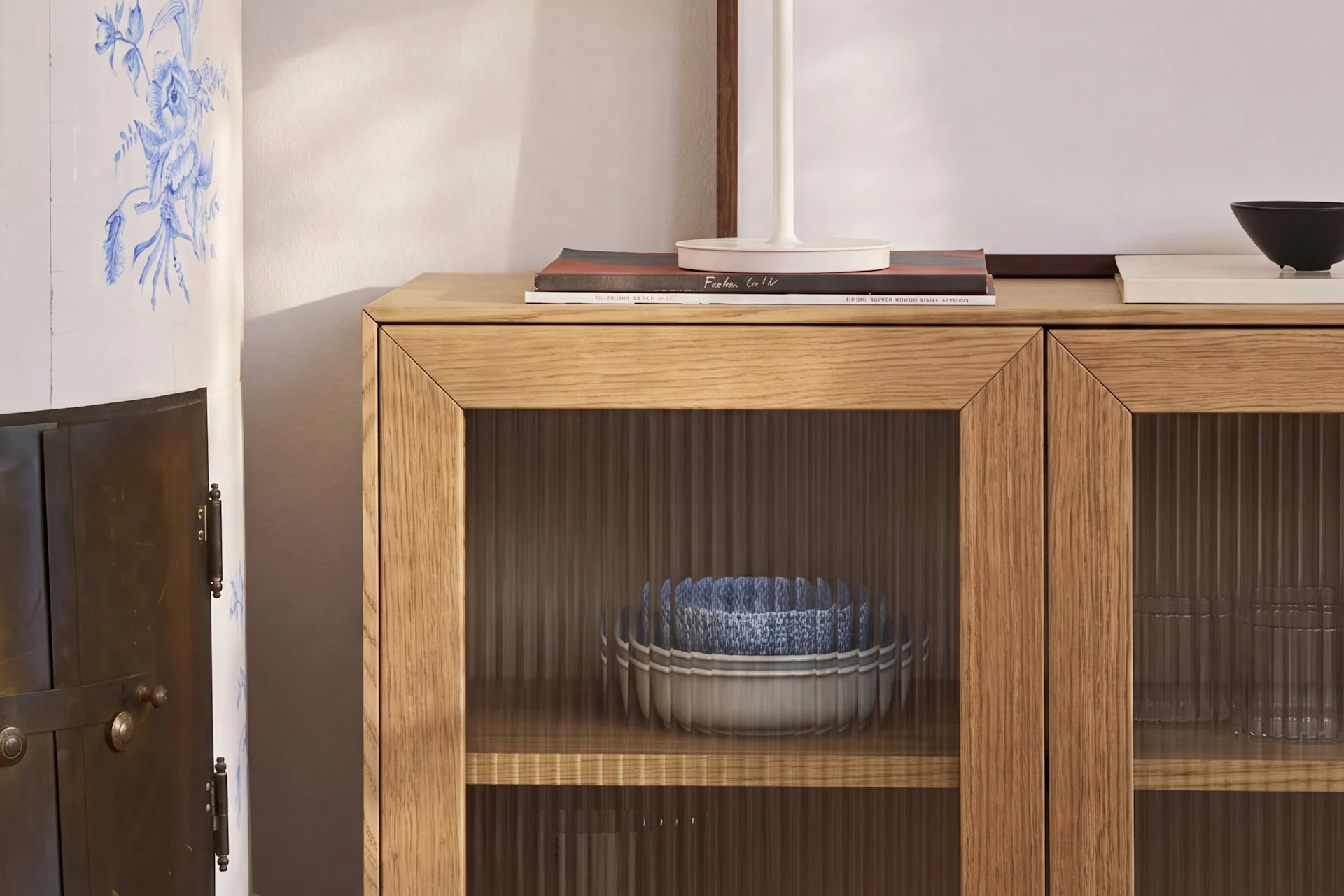 Un armario de madera clara con puertas de cristal estriado contiene cuencos azules y blancos, junto a una estufa de cerámica con motivos florales azules.