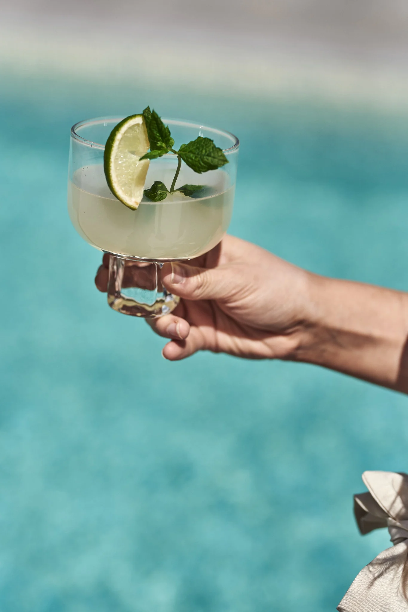Descubre estos refrescantes y sencillos cócteles y mocktails veraniegos. Aquí se ve una mano sosteniendo un mojito con un gajo de lima junto a una piscina.