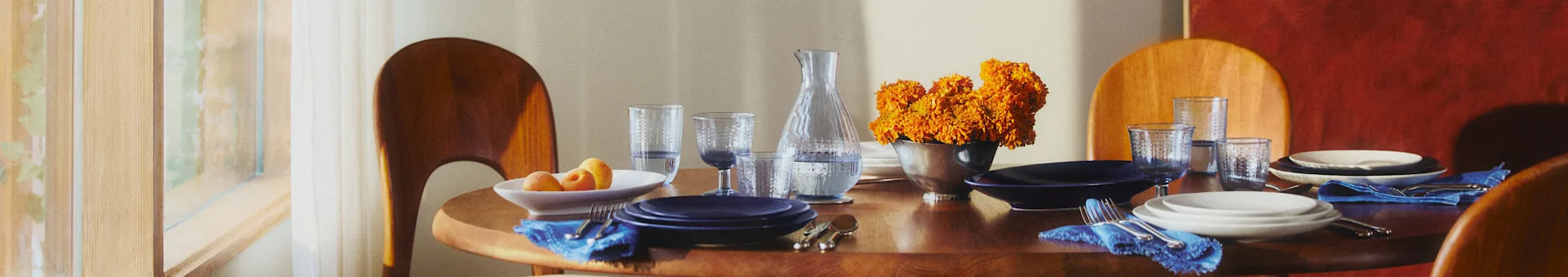 Mesa de comedor redonda de madera con platos azules y blancos, servilletas azules y flores naranjas. La luz del sol se filtra por una ventana.