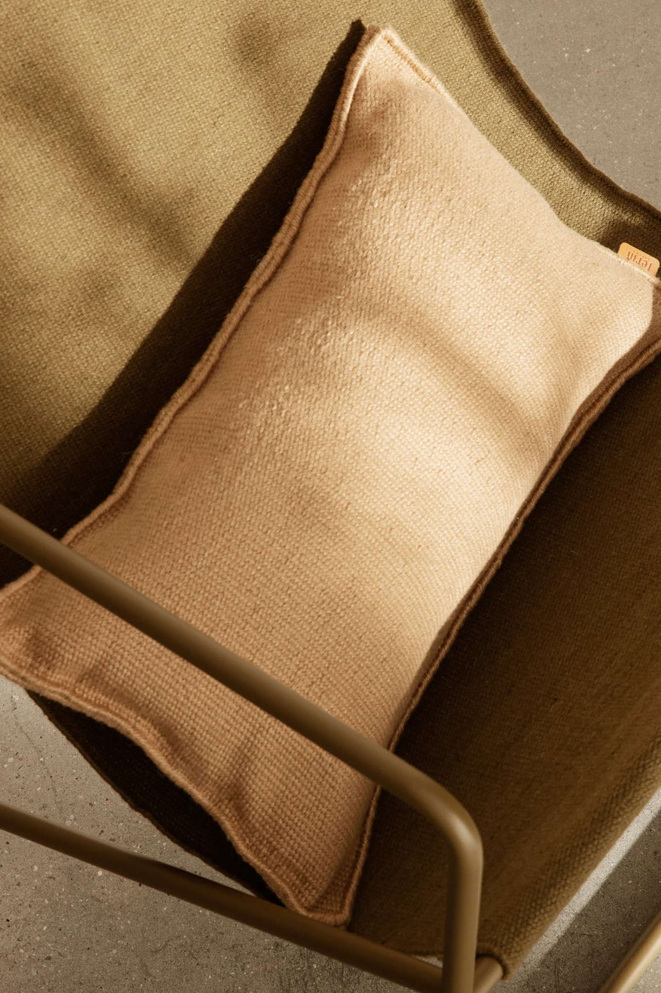 Vista aérea de un cojín marrón claro sobre una silla de tela texturizada verde oliva oscuro, con parte de un marco metálico y suelo de hormigón.