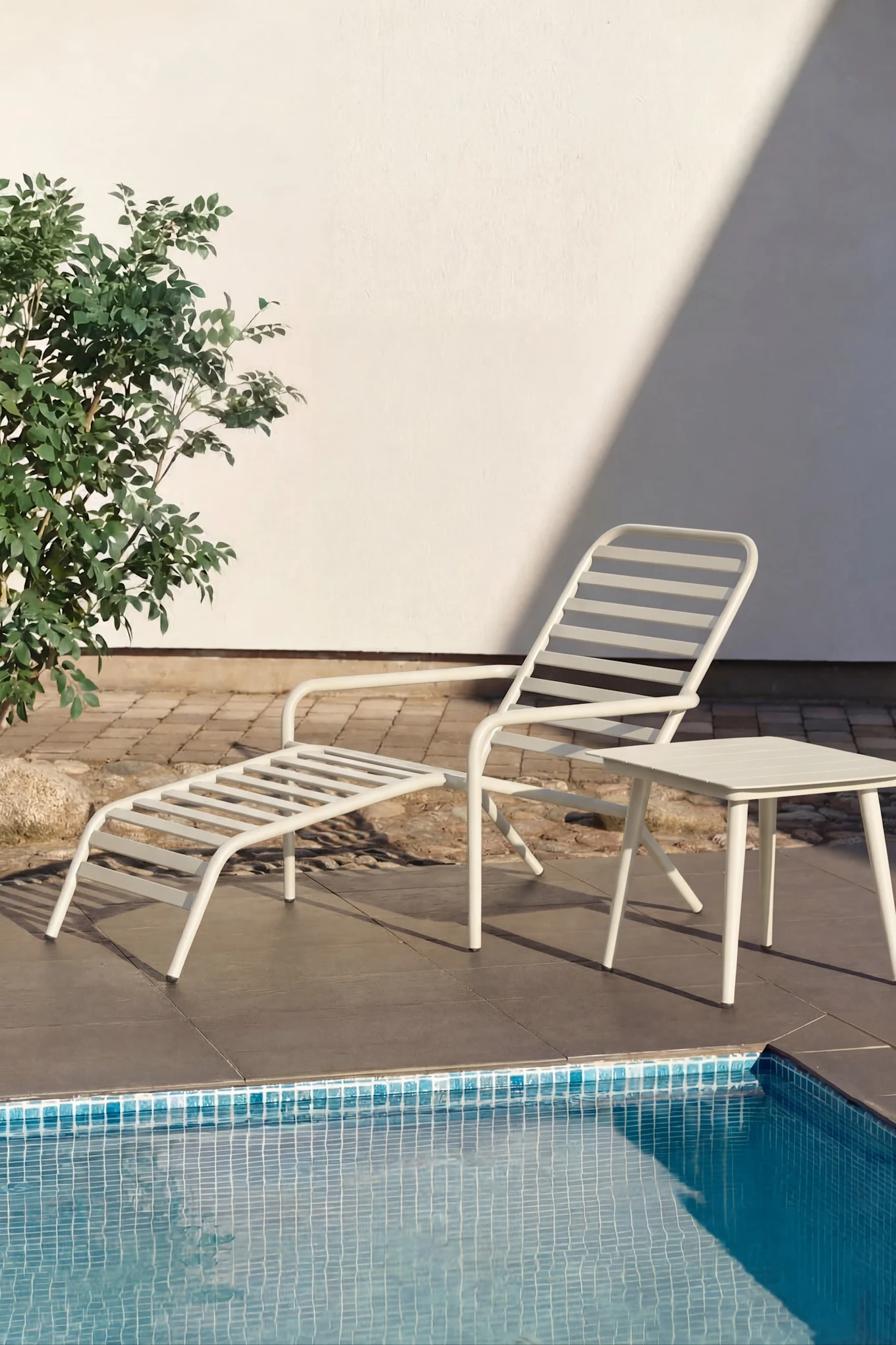 Tumbona blanca y pequeña mesa auxiliar blanca junto a una piscina con agua turquesa. Plantas verdes y una pared blanca al fondo.