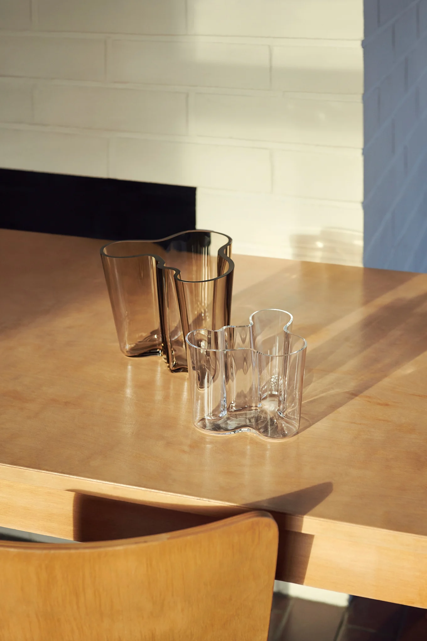 Tres jarrones de cristal ondulados sobre una mesa de madera. Dos son ámbar, uno es transparente, con luz solar y sombras en la escena.
