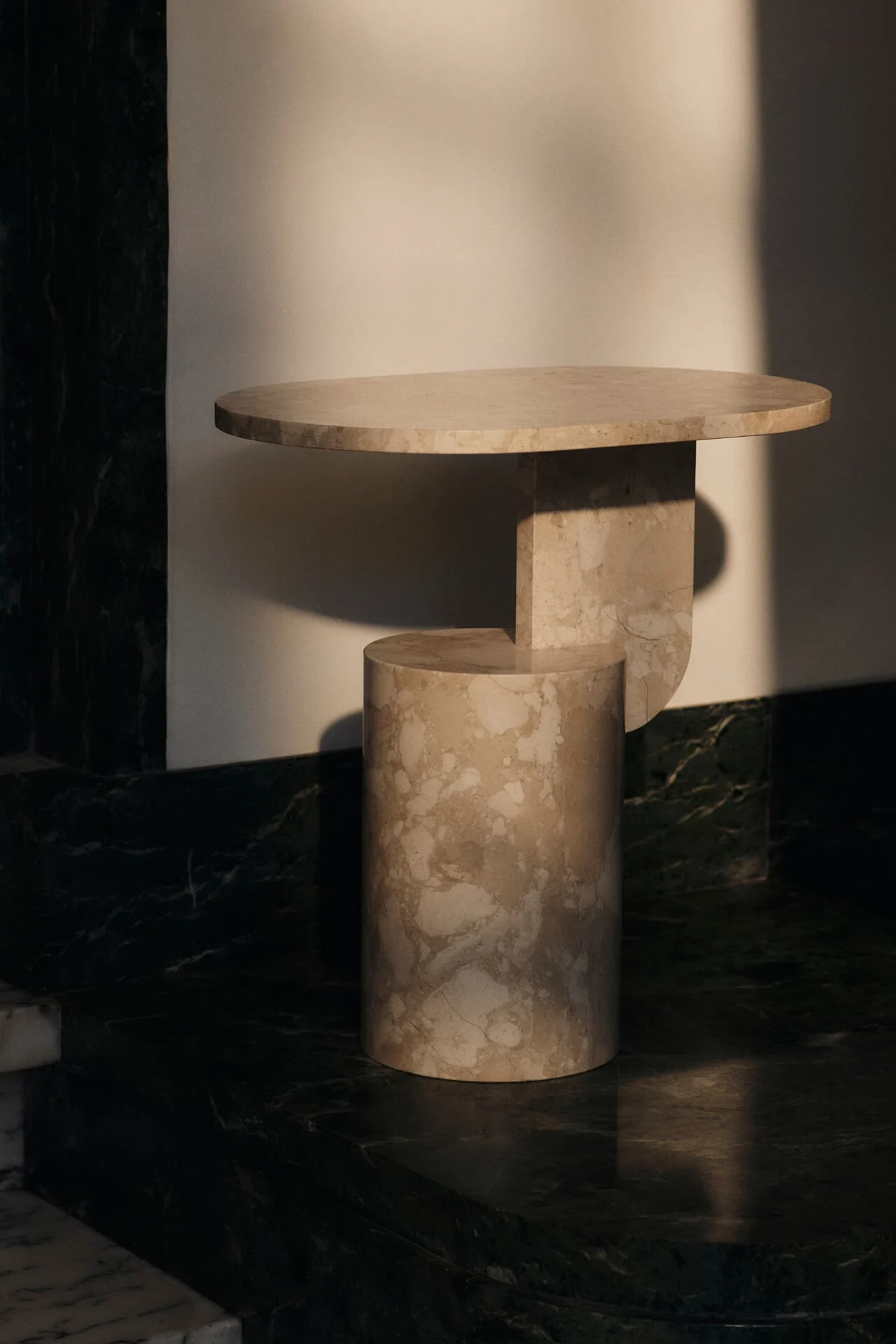 Mesa auxiliar moderna de mármol beige con tapa redonda y base geométrica sobre una pared clara.