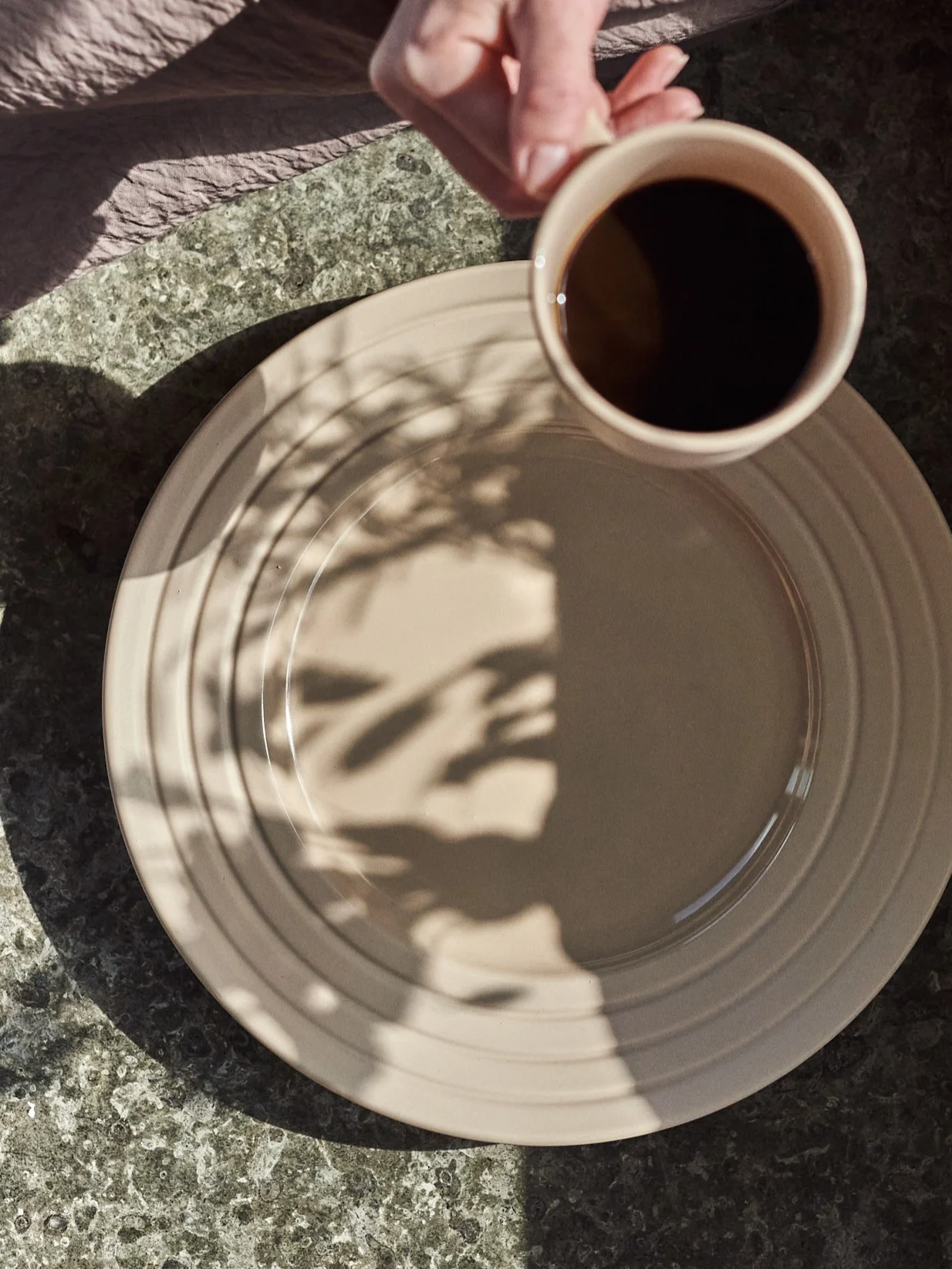 Mano sostiene taza de café sobre plato beige con sombras de hojas en una mesa moteada.