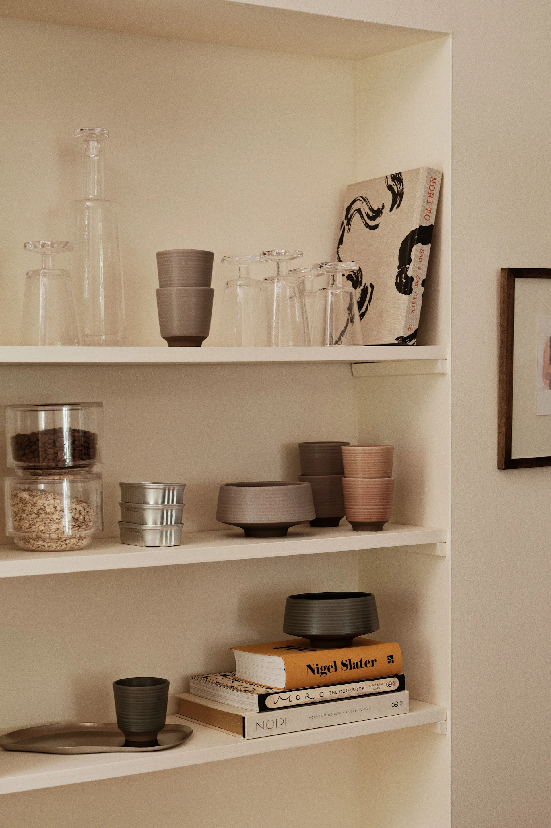 Estantes empotrados exhiben cristalería, tazas y cuencos de cerámica apilados en tonos neutros, recipientes de comida, latas plateadas y libros de cocina.