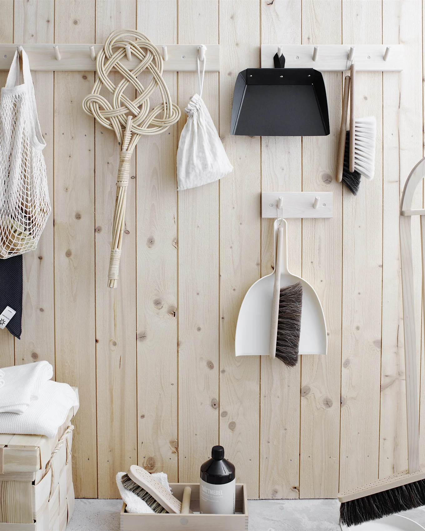 Herramientas de limpieza y artículos de lavandería organizados en una pared de madera clara. Incluye cepillos, recogedores, un batidor de alfombras, una bolsa de red, toallas y detergente.