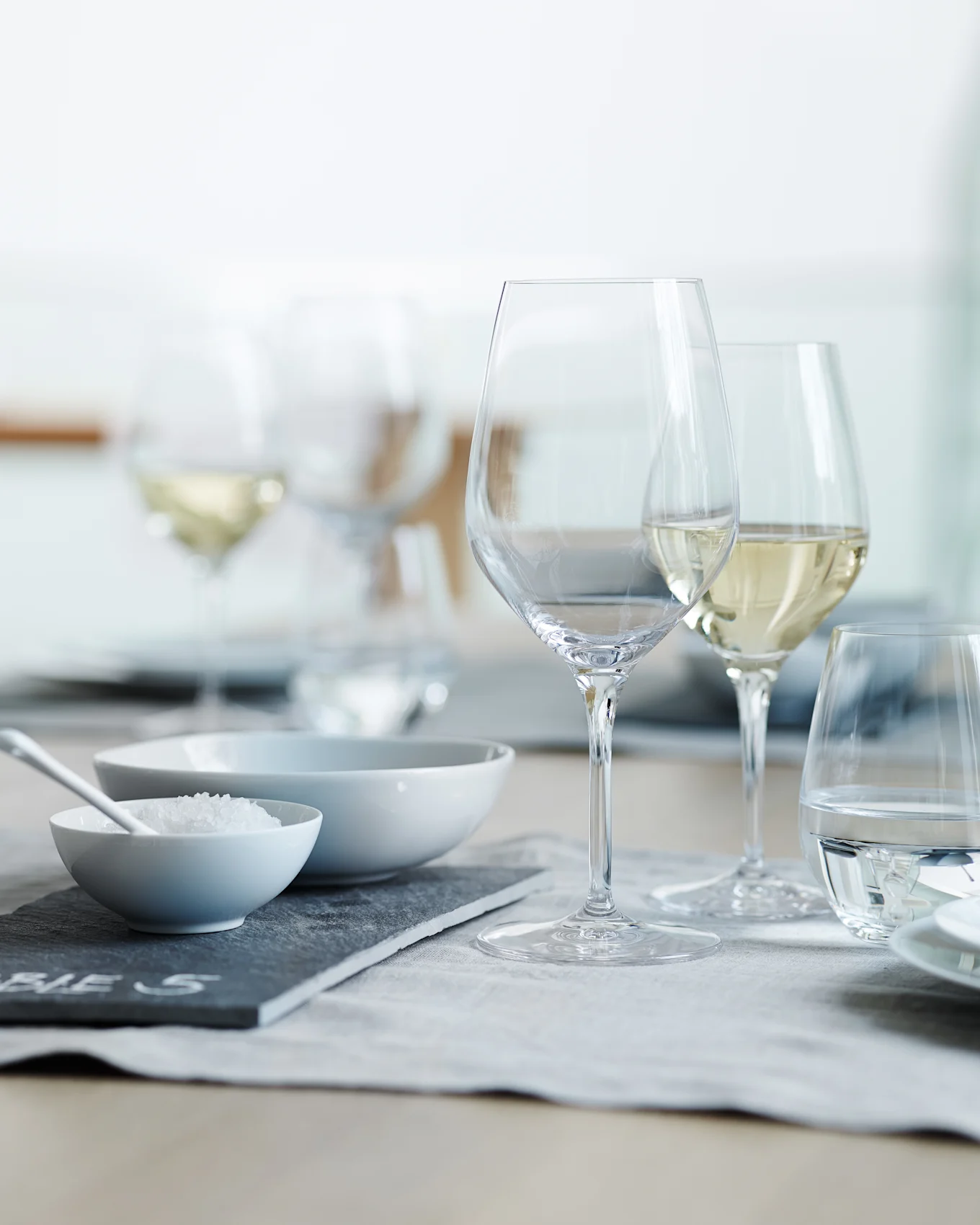 Una mesa de comedor puesta con copas de vino y vasos de agua transparentes, un cuenco con sal gruesa y un cuenco vacío sobre un mantel individual gris.