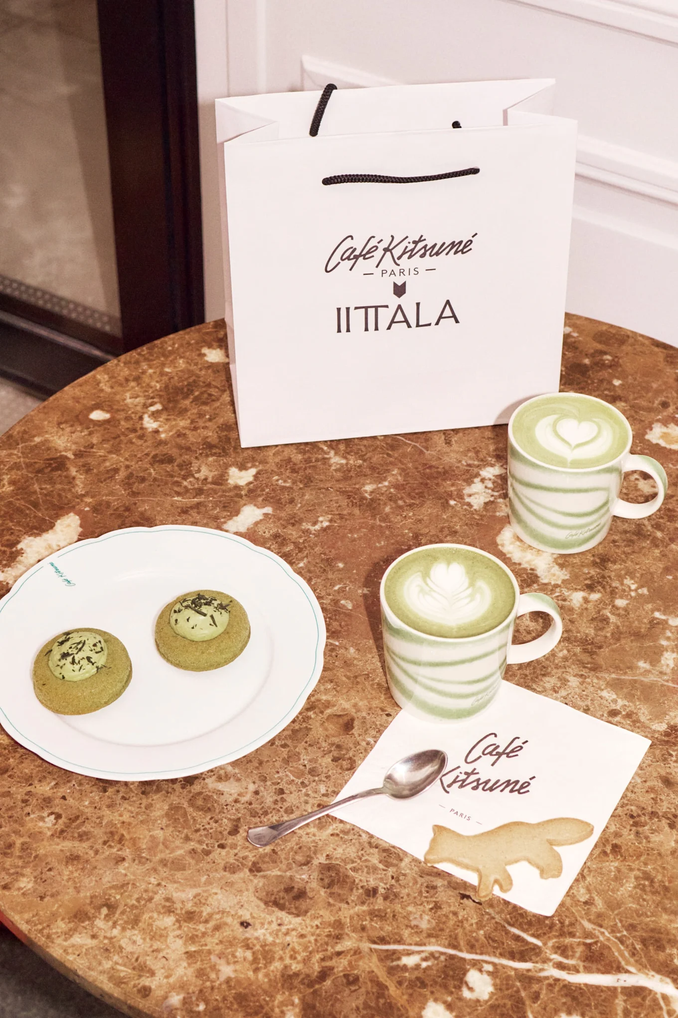 Una bolsa de compras blanca, dos lattes matcha, dos pasteles verdes, una galleta de zorro y una servilleta sobre una mesa de mármol.