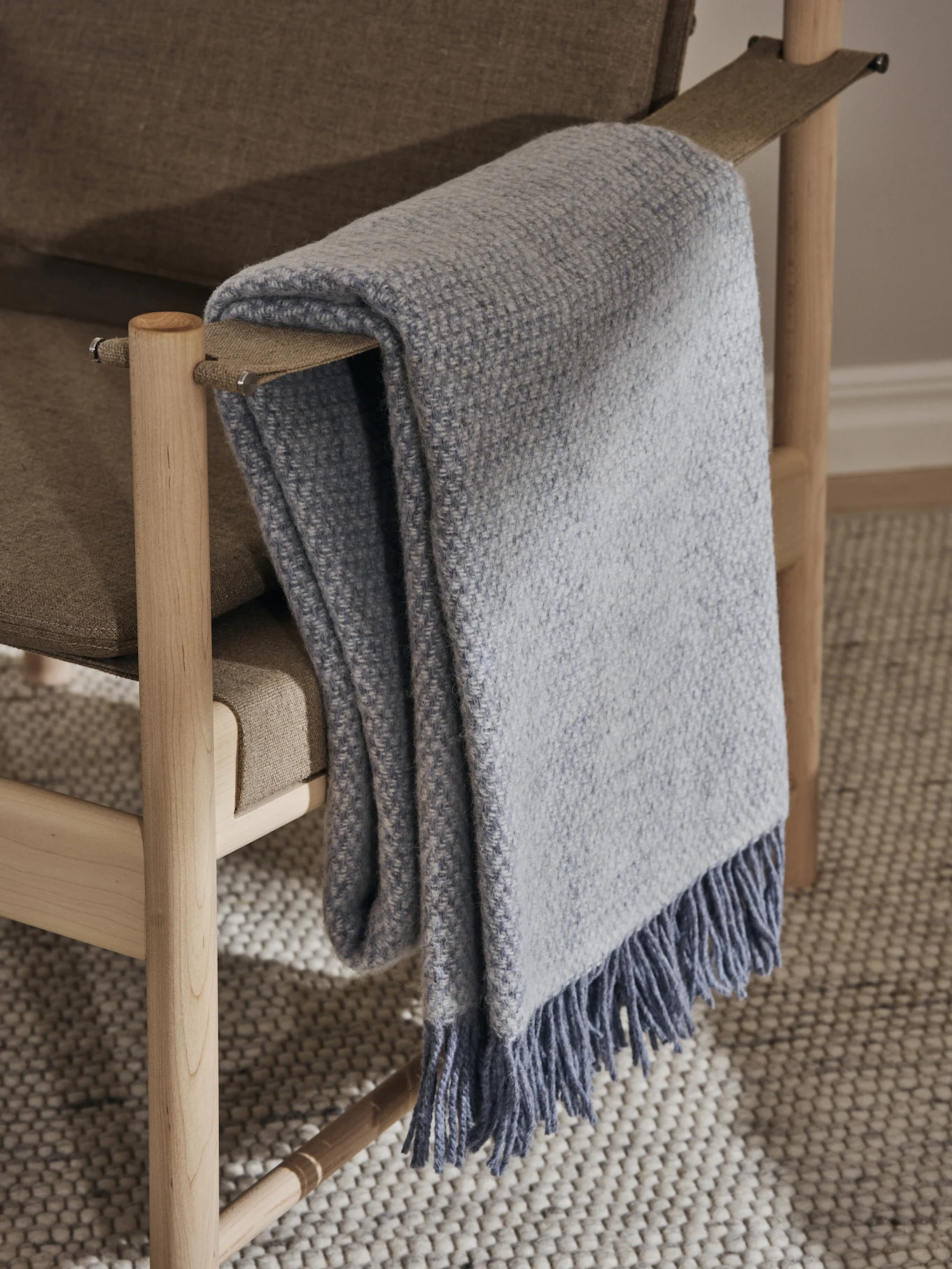 Manta tejida azul claro con flecos colocada sobre una silla tapizada de color marrón claro con estructura de madera clara, sobre una alfombra texturizada.