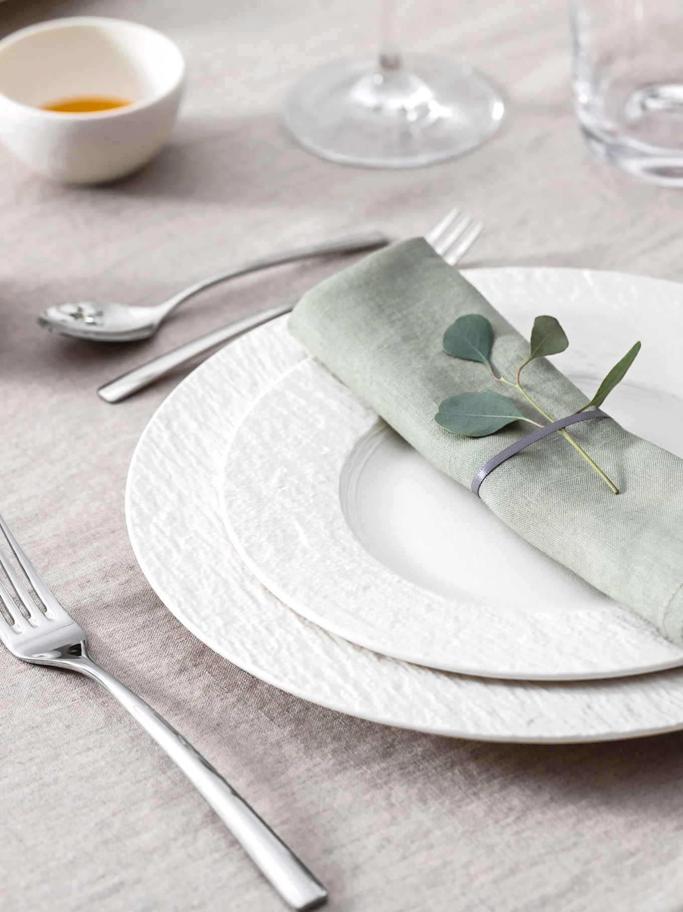 Elegante mesa puesta con servilleta de lino verde claro, hojas de eucalipto, platos blancos texturizados y cubiertos sobre un mantel beige.