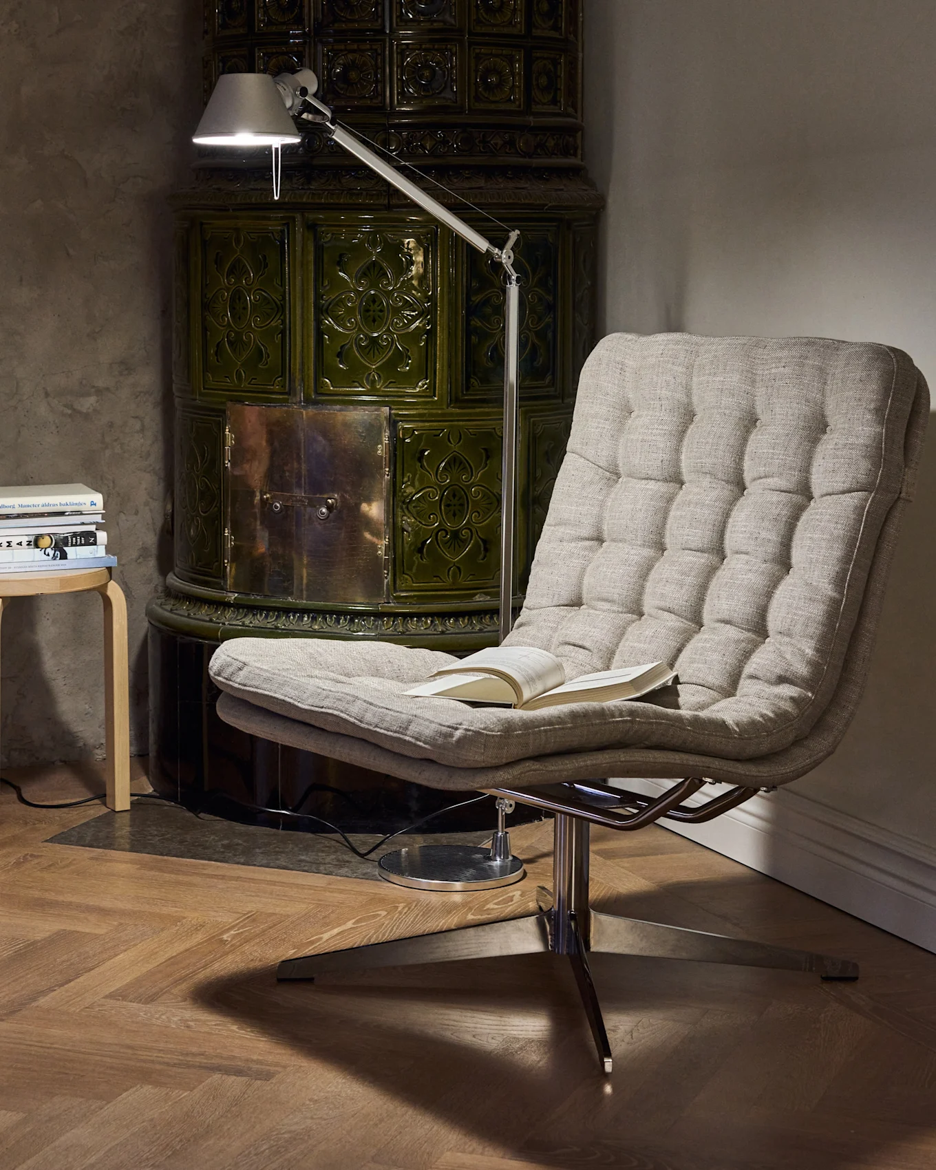 Acogedor rincón de lectura con un sillón capitoné, una lámpara de pie ajustable y una estufa de azulejos verde oscuro.