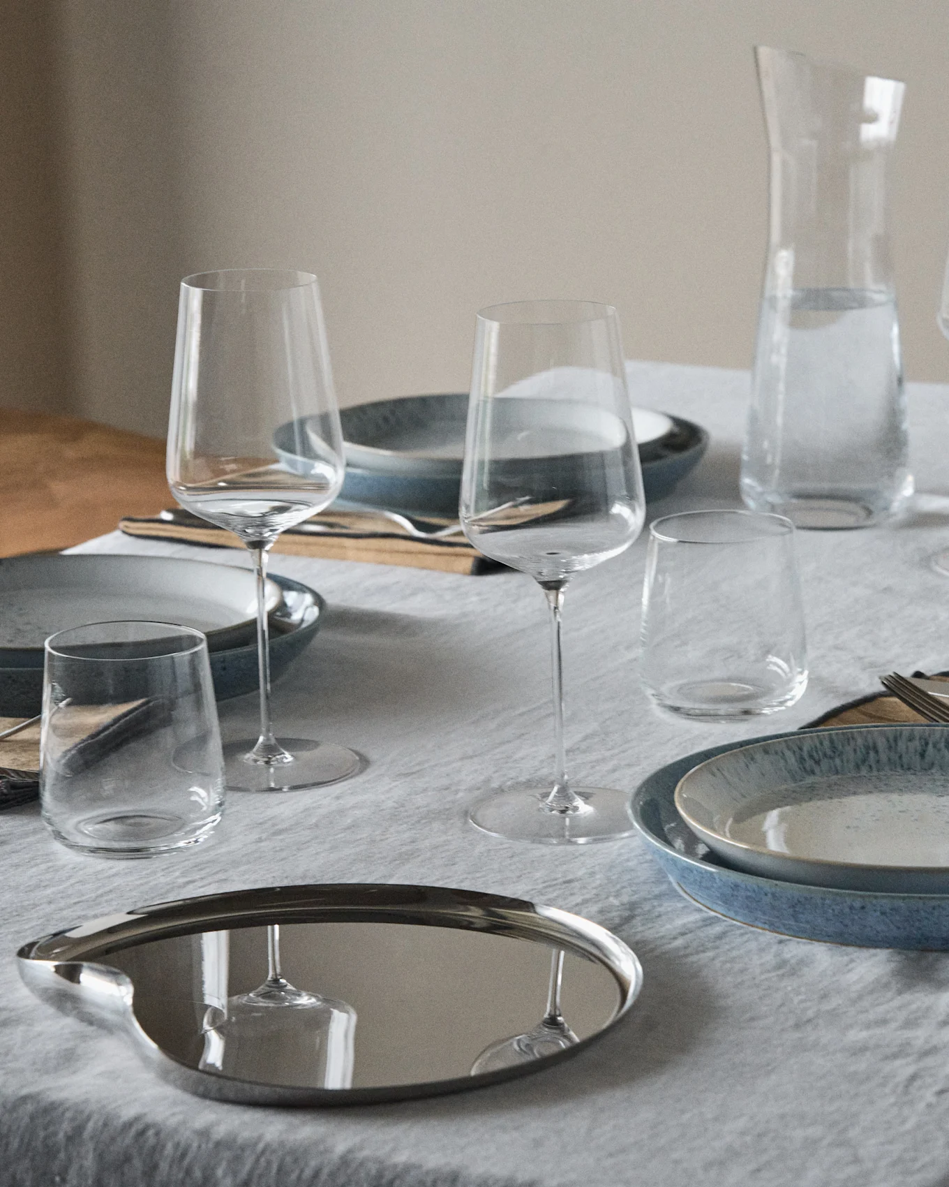 Mesa puesta con platos azules y moteados, copas de vino, vasos, una jarra de agua y una bandeja ovalada reflectante.